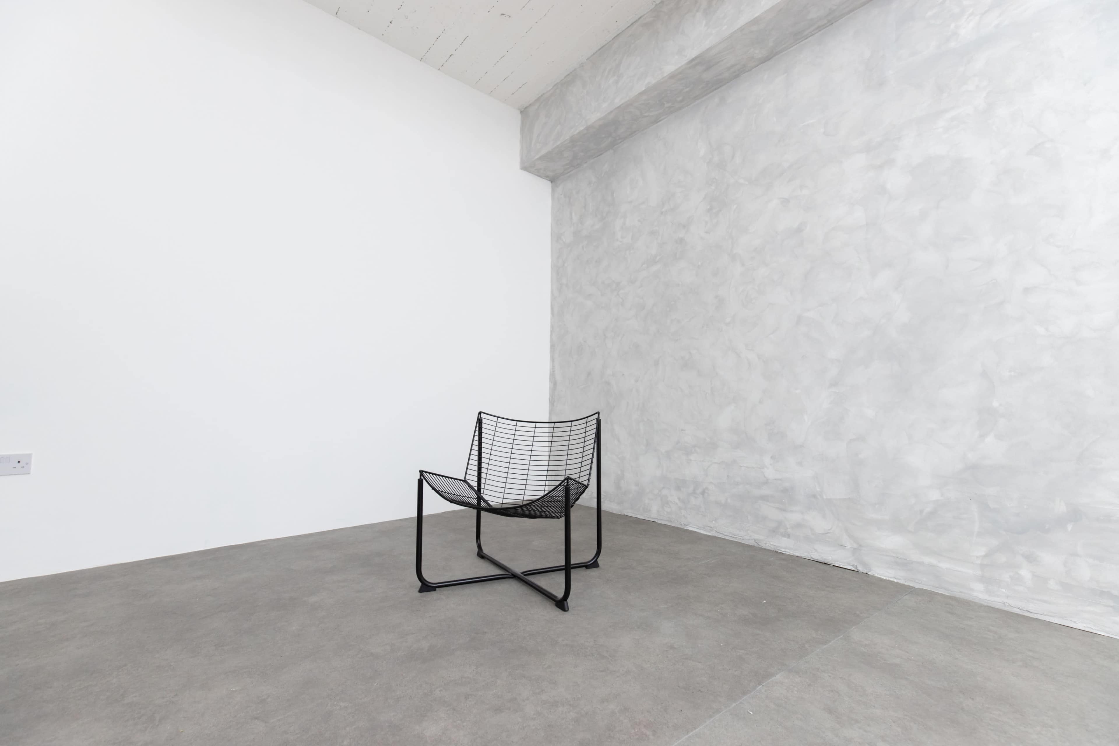 A minimalist room features a black wire chair against a plain white wall and a textured gray wall.