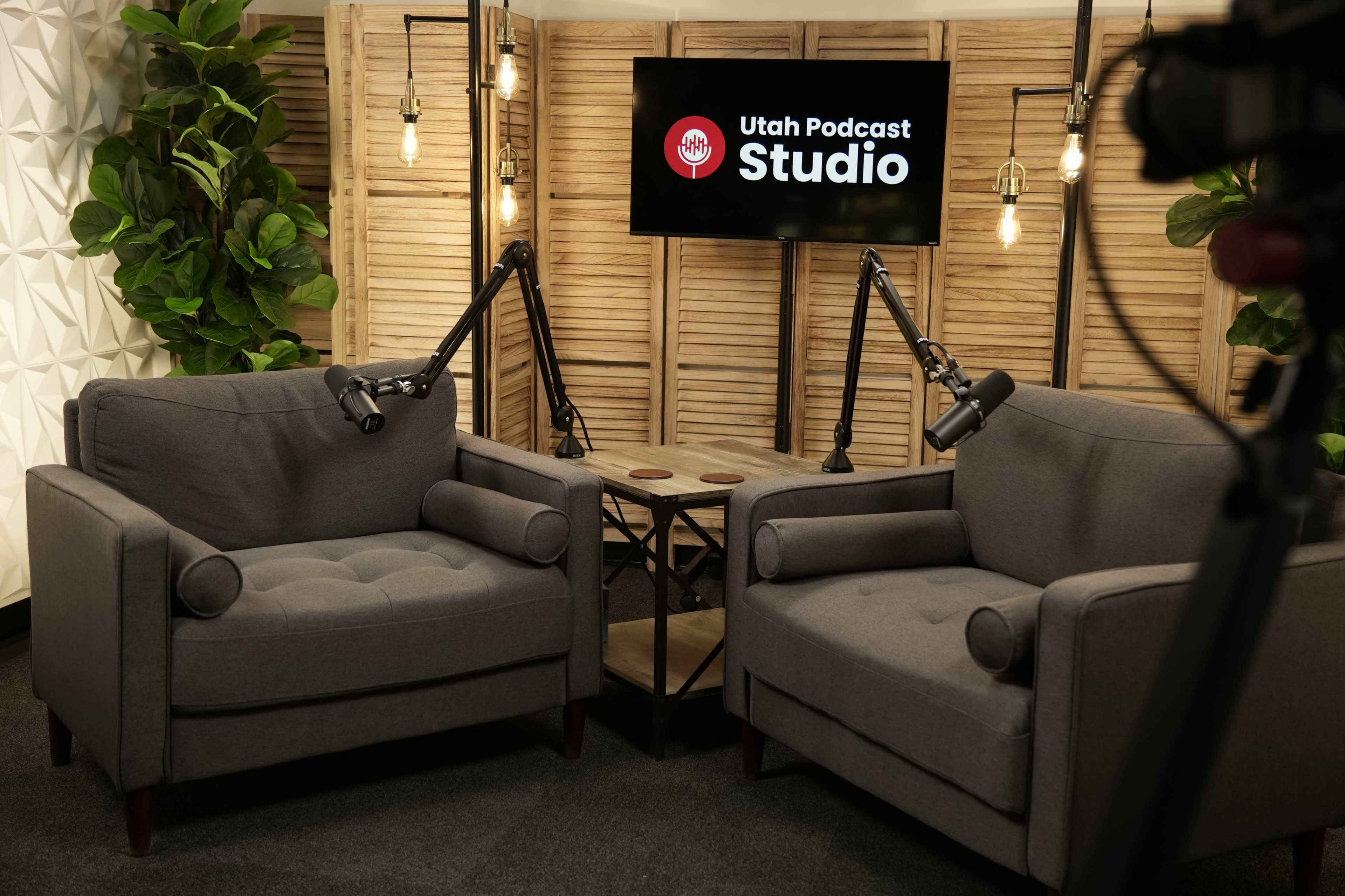 The image shows a podcast studio setting with two gray armchairs, adjustable microphones, and a backdrop featuring a logo that reads "Utah Podcast Studio."
