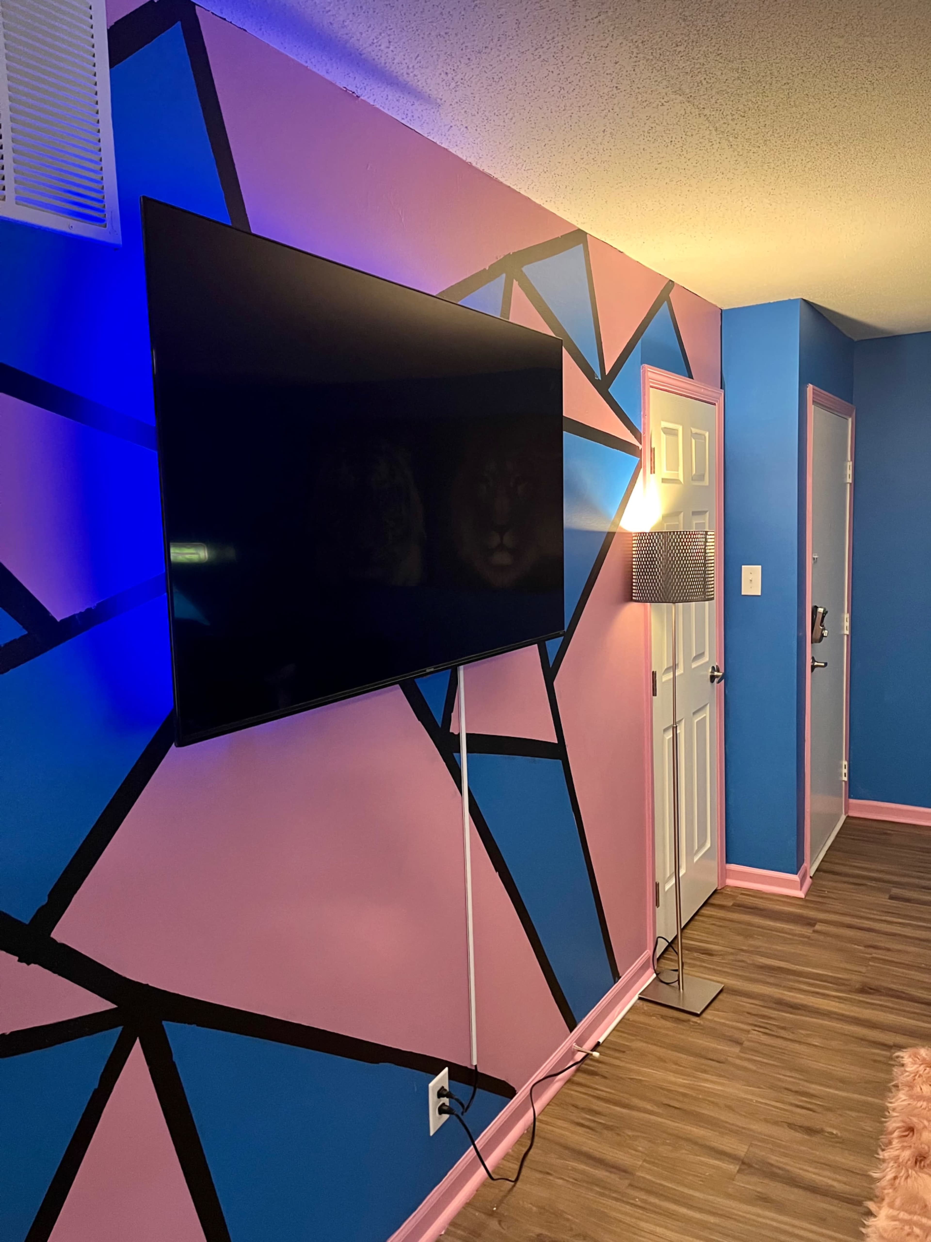 A modern living space with a large flat-screen TV mounted on a wall featuring geometric blue and pink patterns, alongside a sleek floor lamp.