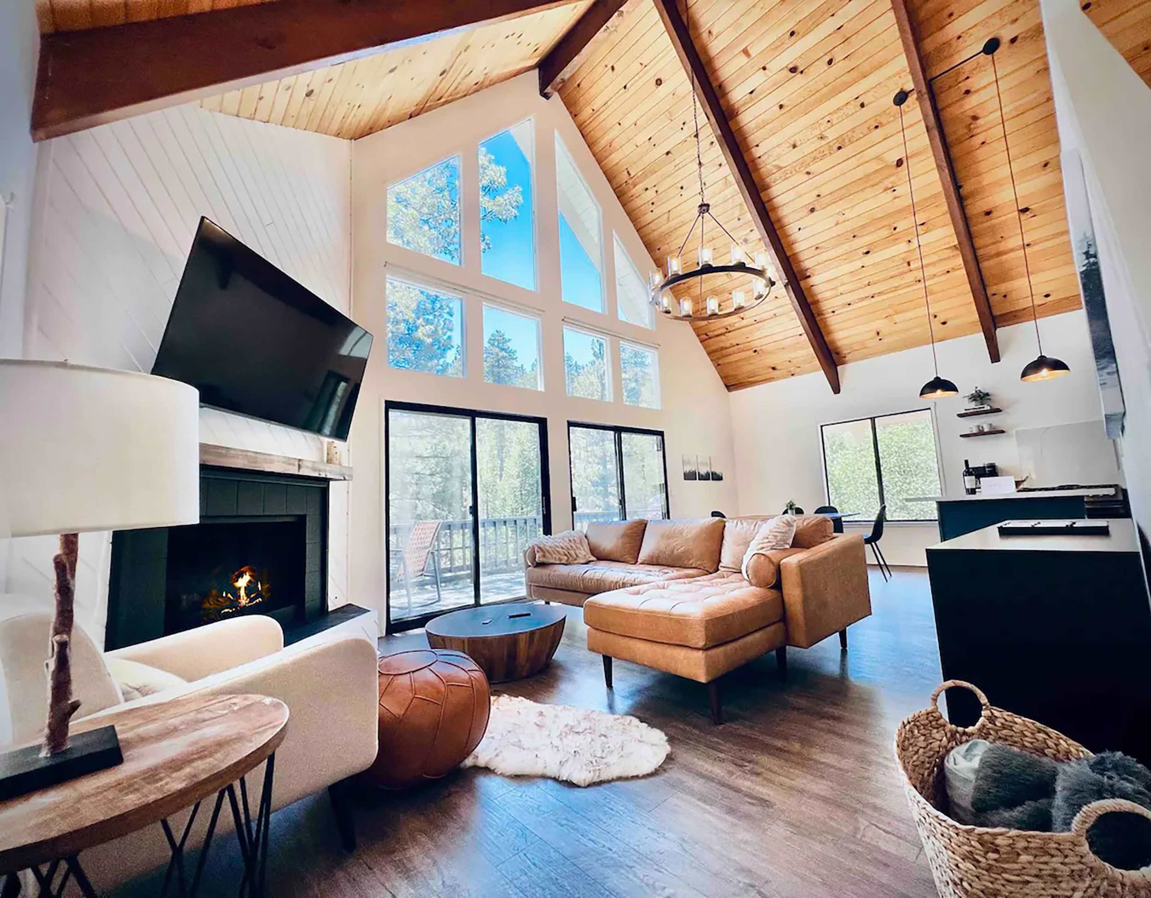 The image shows a spacious, modern living room with high ceilings, large windows, a fireplace, and a combination of comfortable furniture.