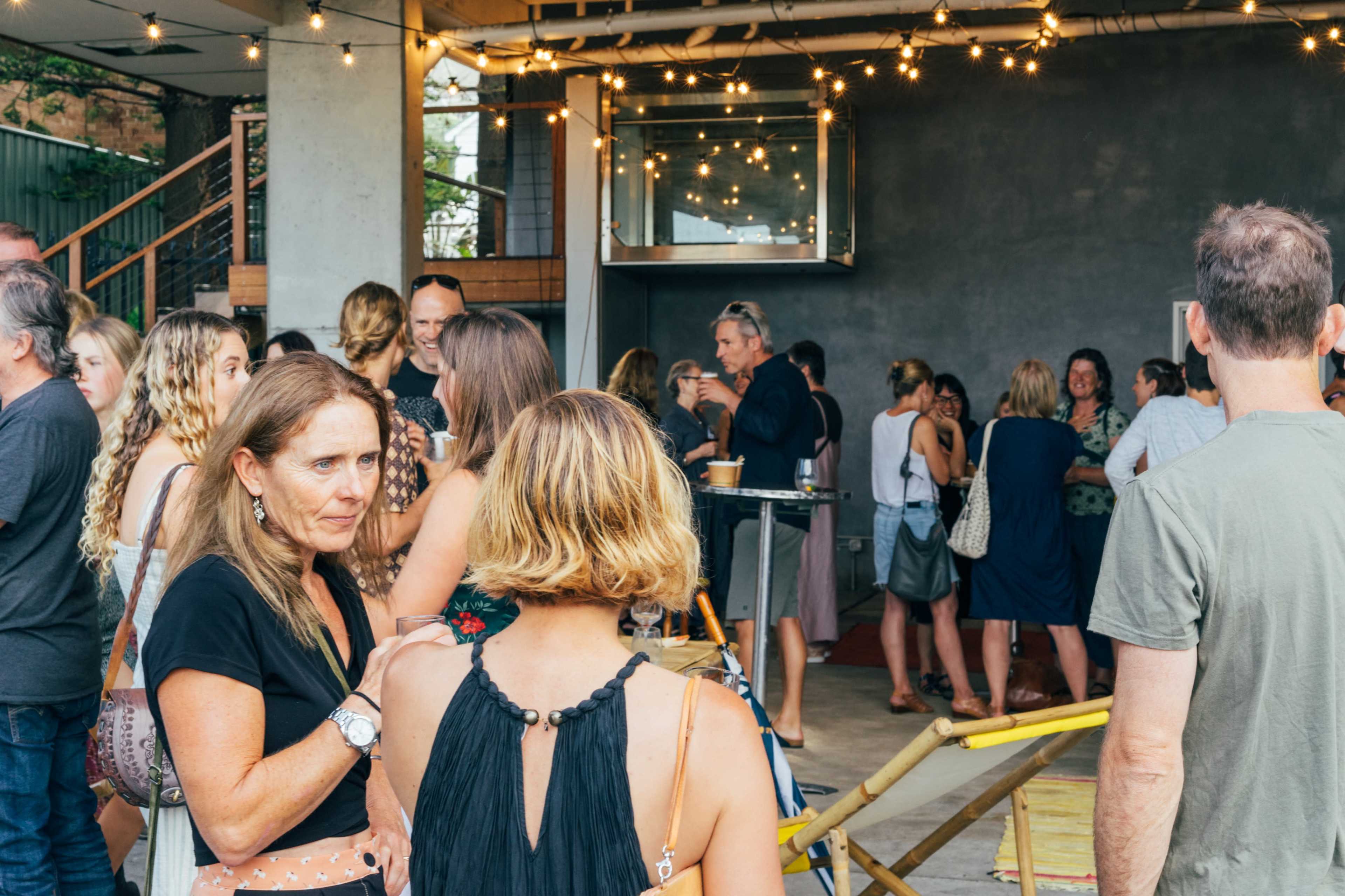 A group of people gathers in a well-lit space, engaging in conversation while surrounded by string lights and a bar area.