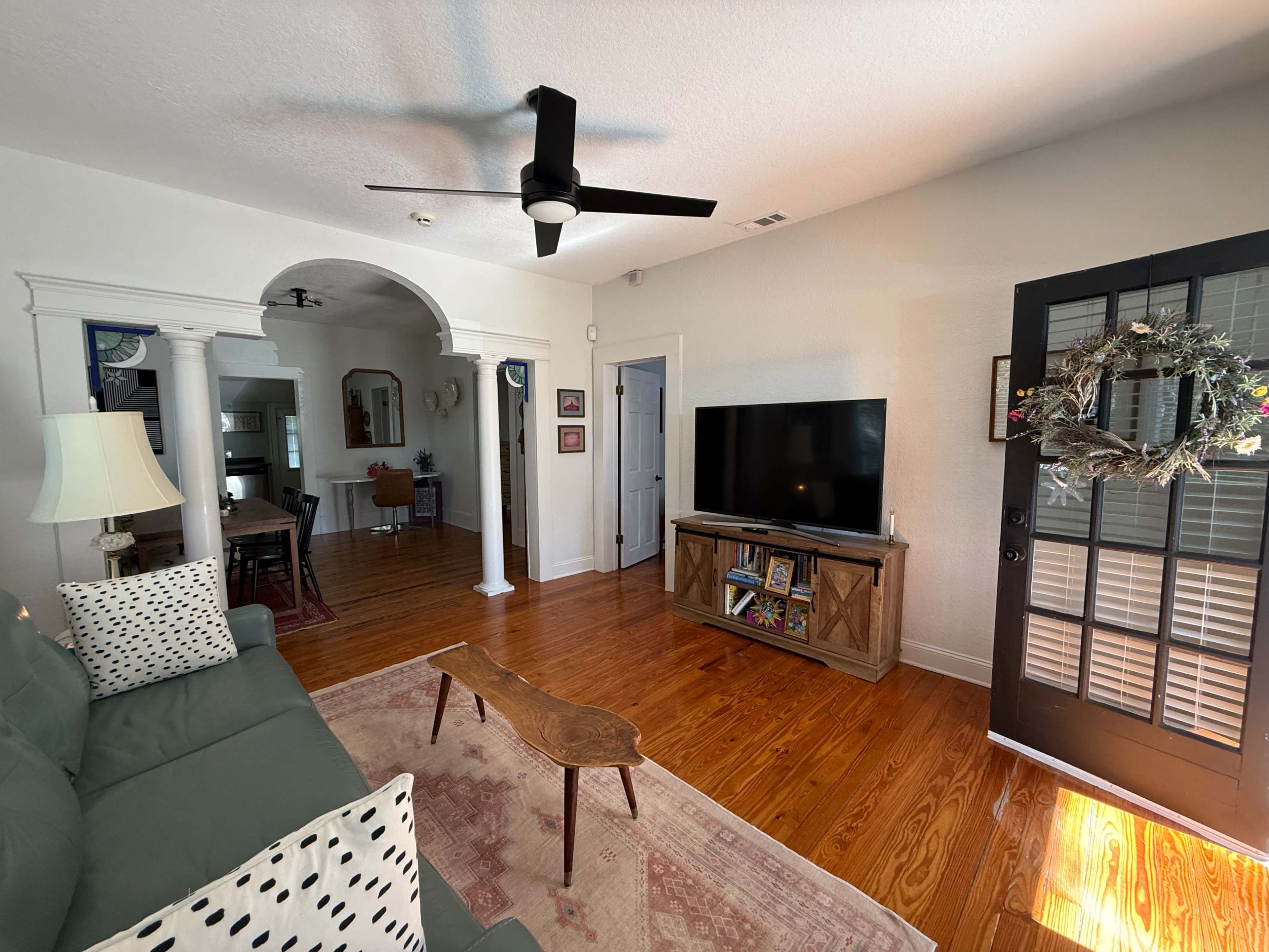 The image shows a cozy living room with a green couch, wooden coffee table, and a television on a stand, next to a doorway leading to another area of the house.