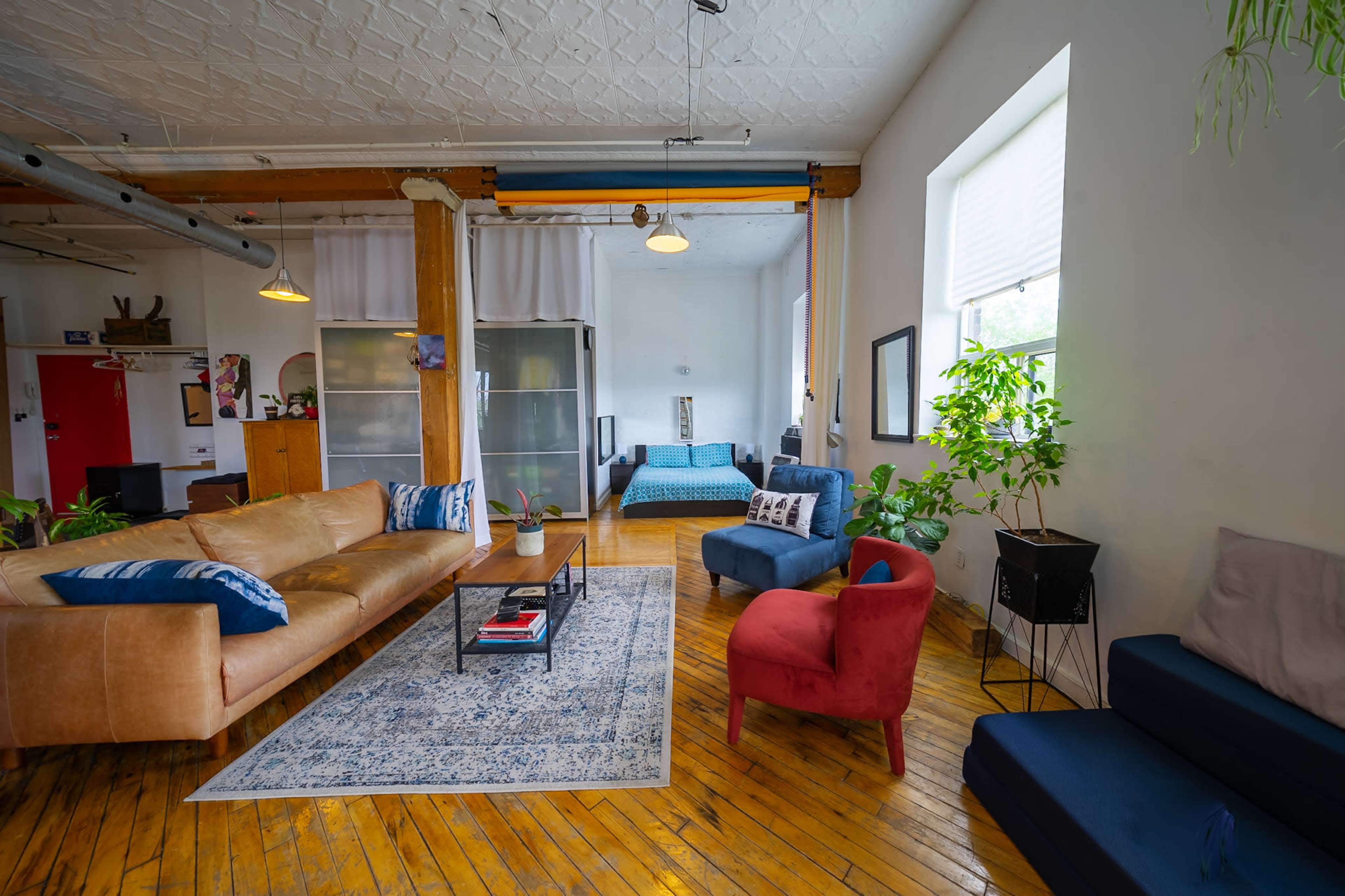 The image depicts a spacious, light-filled apartment with wooden floors, featuring a large brown sofa, a blue chair, and a red chair, along with a coffee table and various plants.