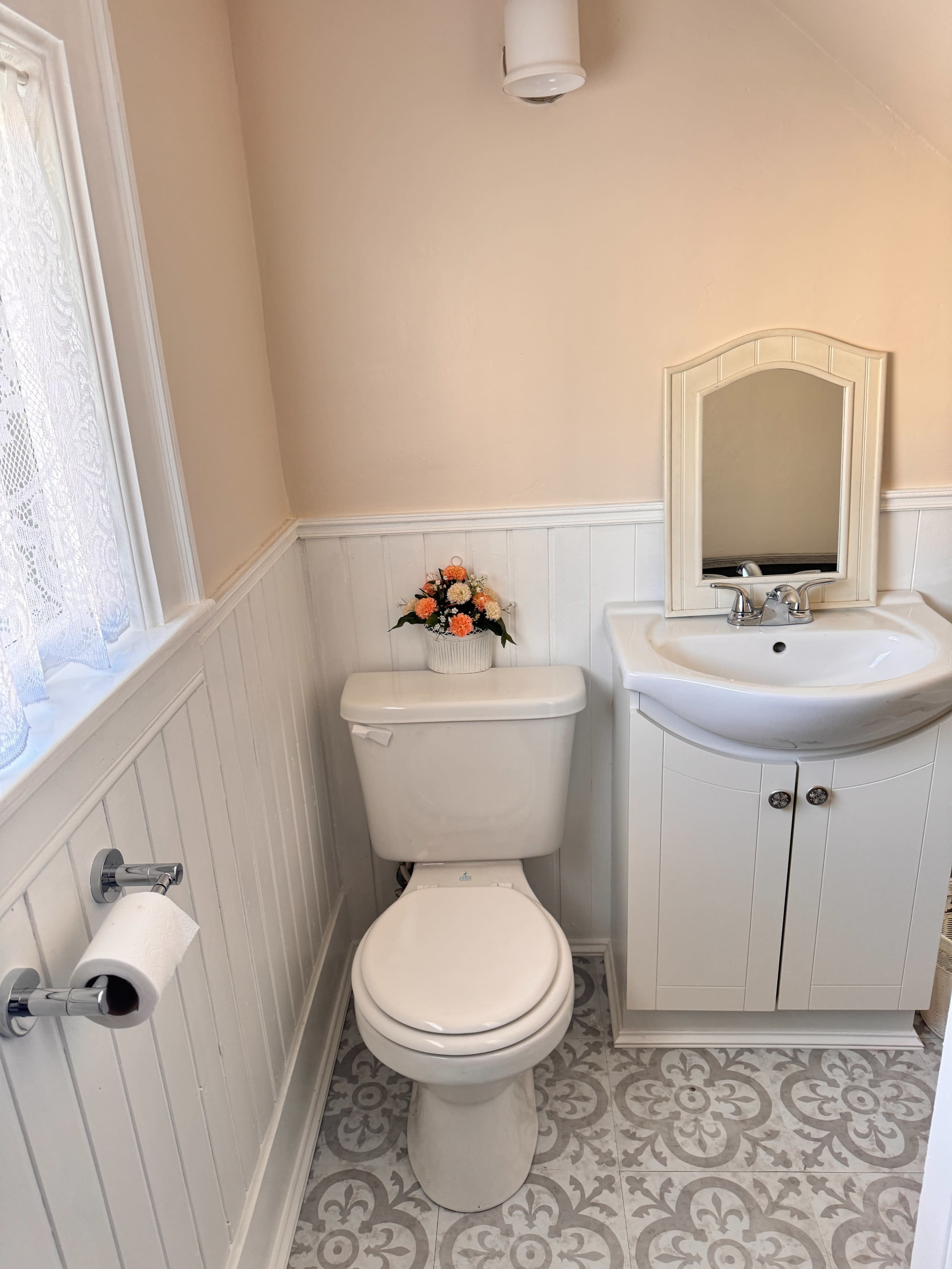 The image shows a small bathroom featuring a toilet, a sink with a mirror, and a decorative flower arrangement on the sink.