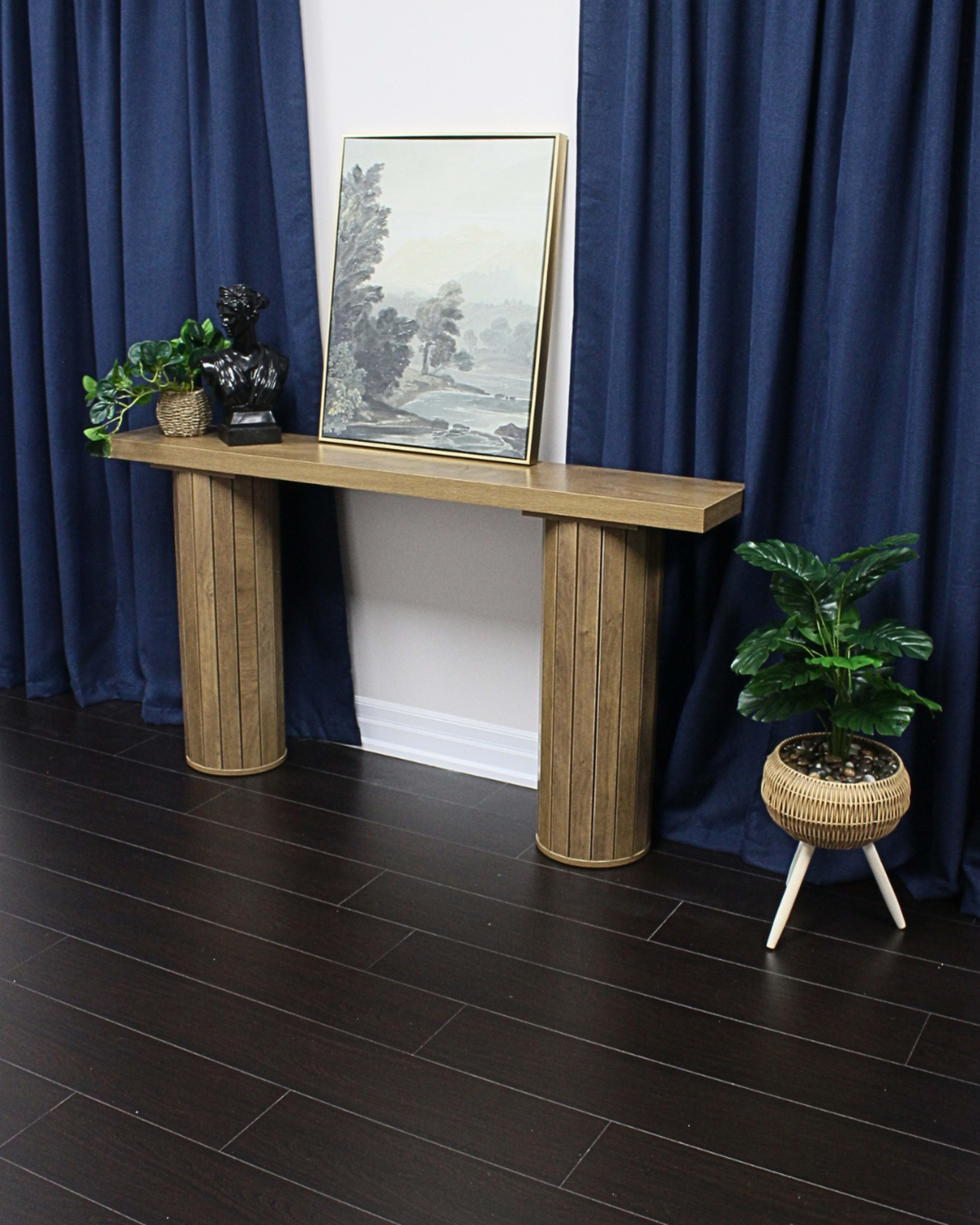 A wooden console table with a framed landscape painting on it is positioned against a wall, flanked by dark blue curtains and potted plants.