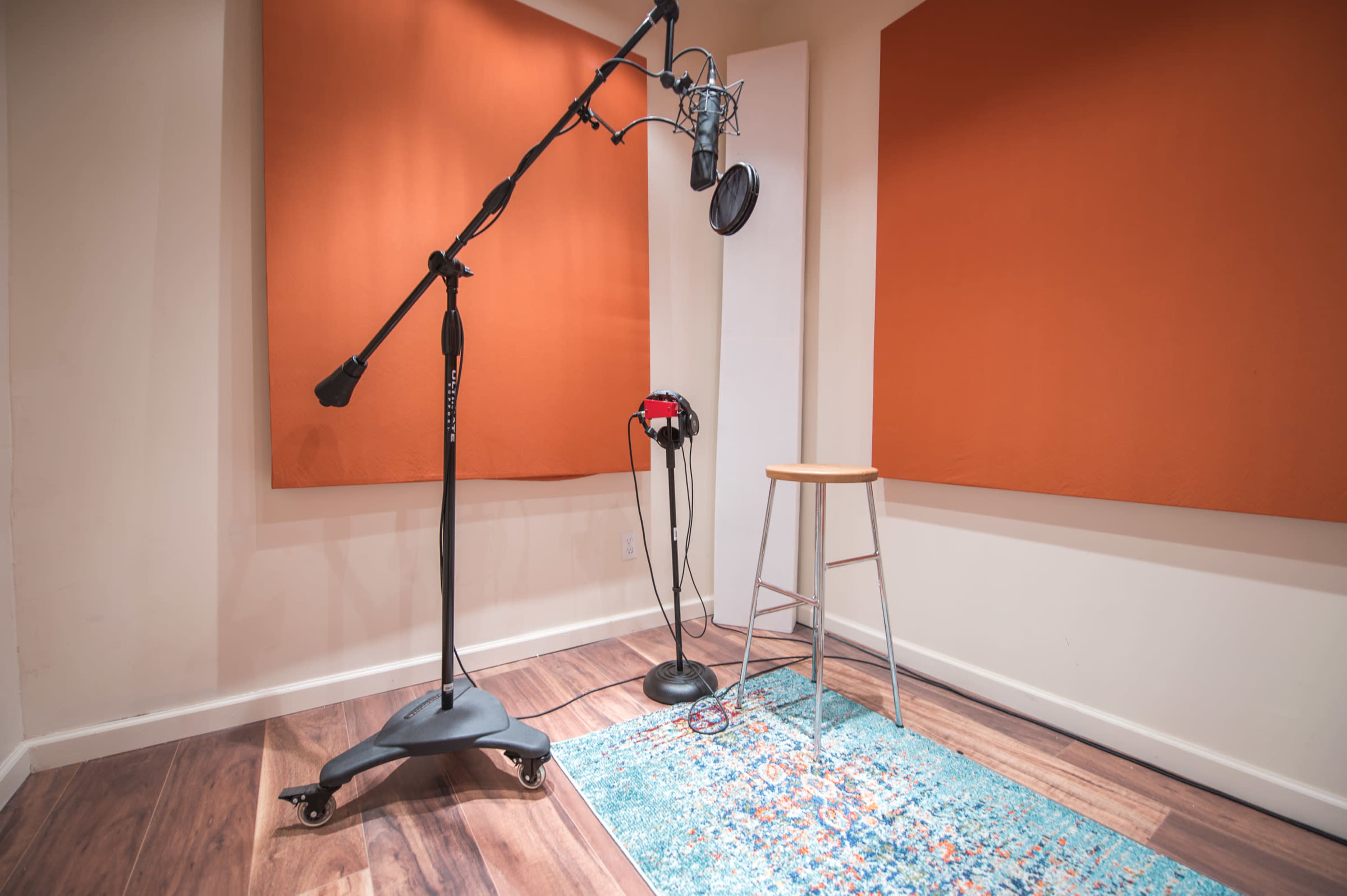 The image shows a small recording studio with a microphone on a stand, headphones, a stool, and soundproofing panels on the walls.