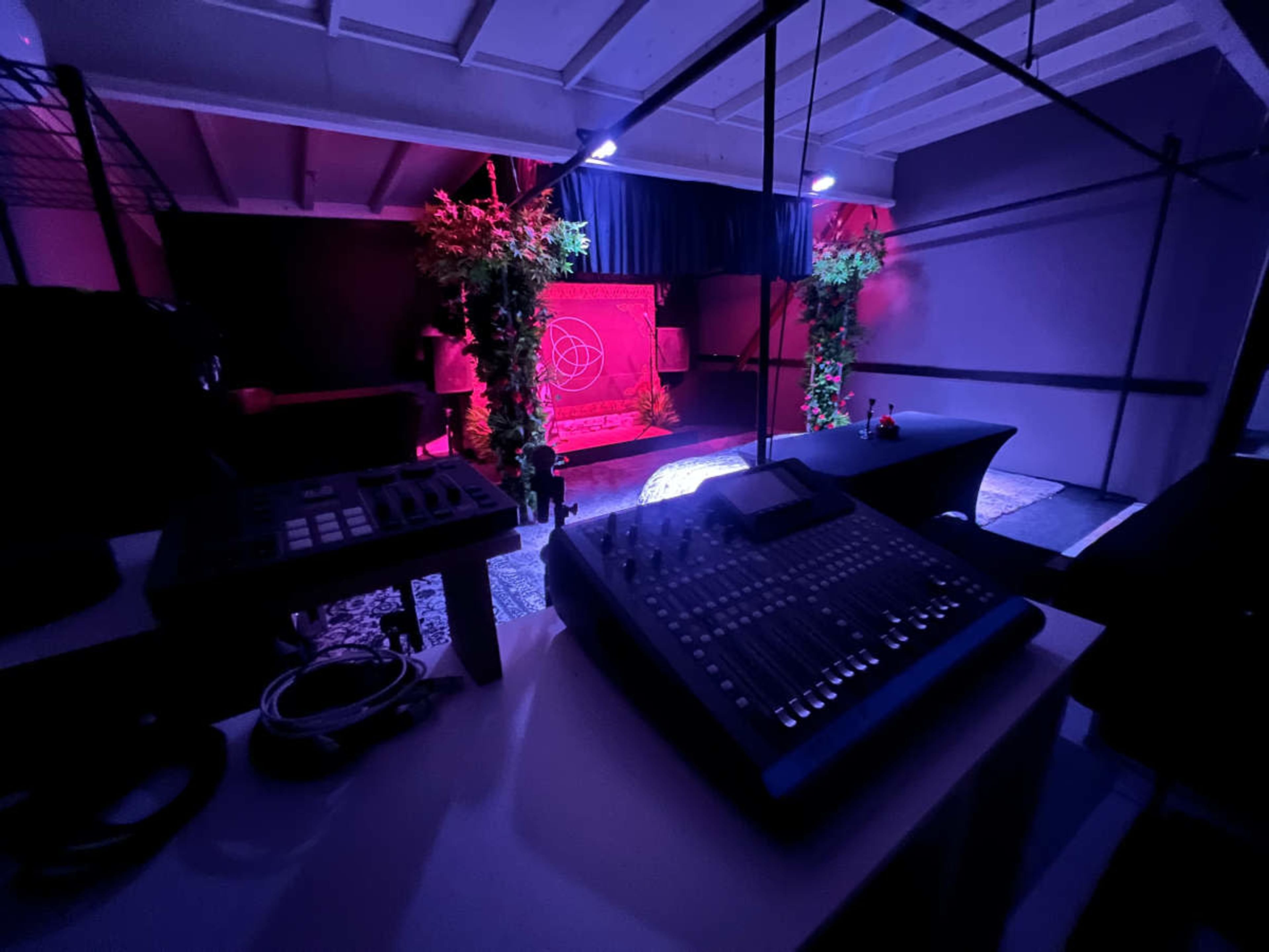 The image shows a stage setup with lighting effects, a sound mixing board in the foreground, and decorative elements like plants in the background.