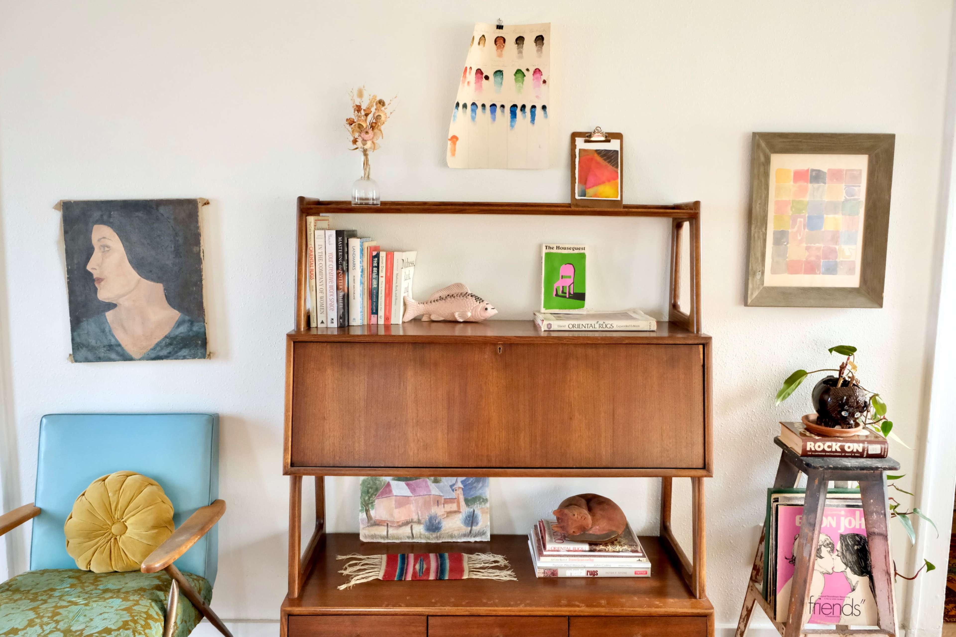 A wooden bookshelf displaying books, decorative items, and artwork, alongside a chair in a soft blue hue.