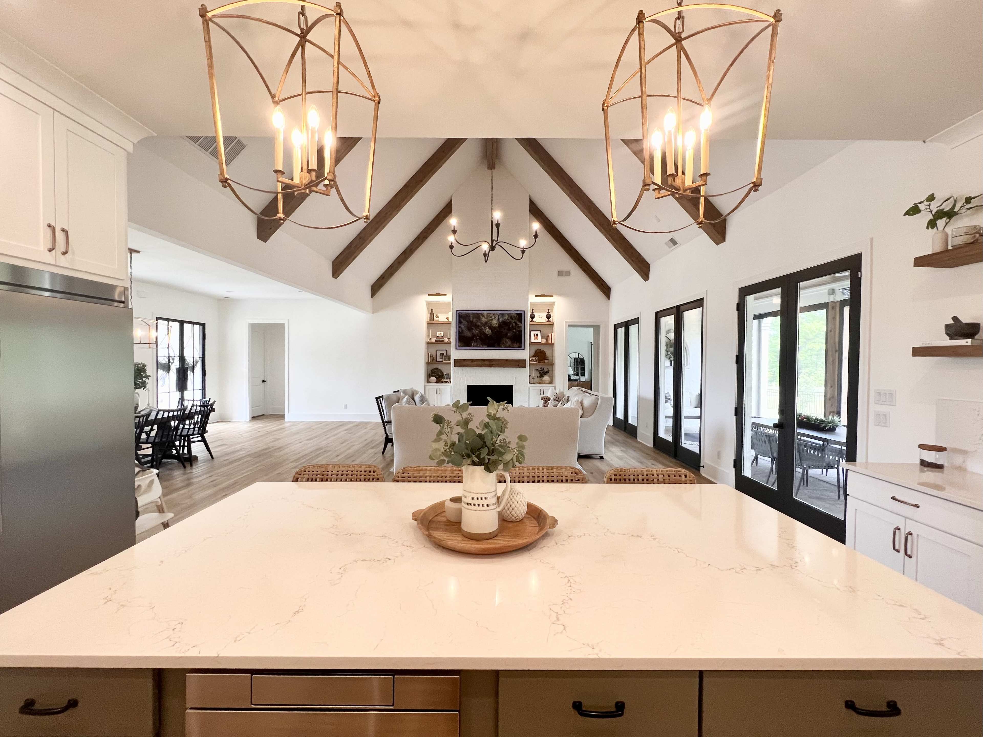 A modern kitchen features a marble island and pendant lights, leading into a spacious living area with a high, beamed ceiling and large windows.