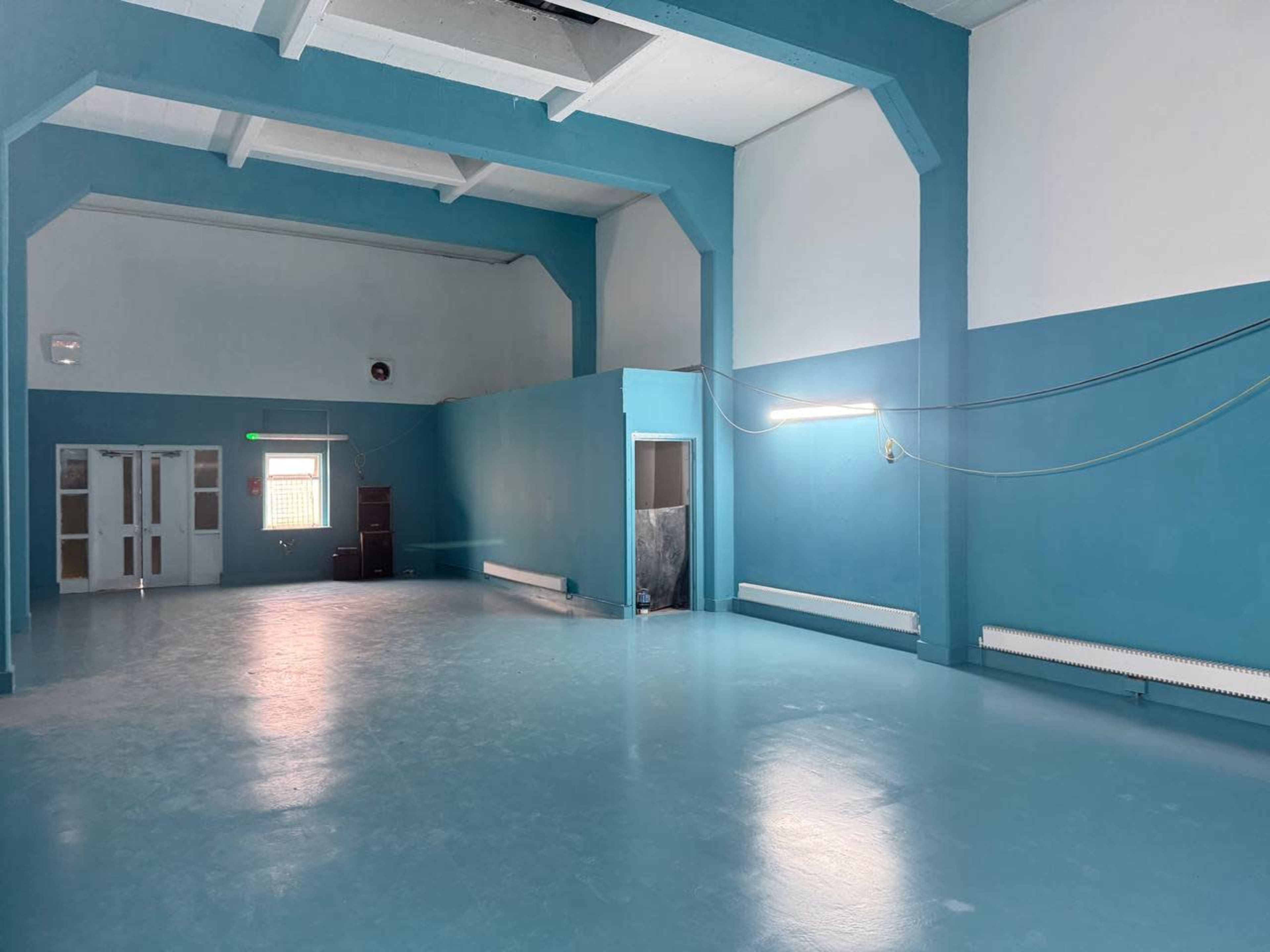 The image shows a spacious room with blue walls and a polished floor, featuring large windows and minimal furnishings.
