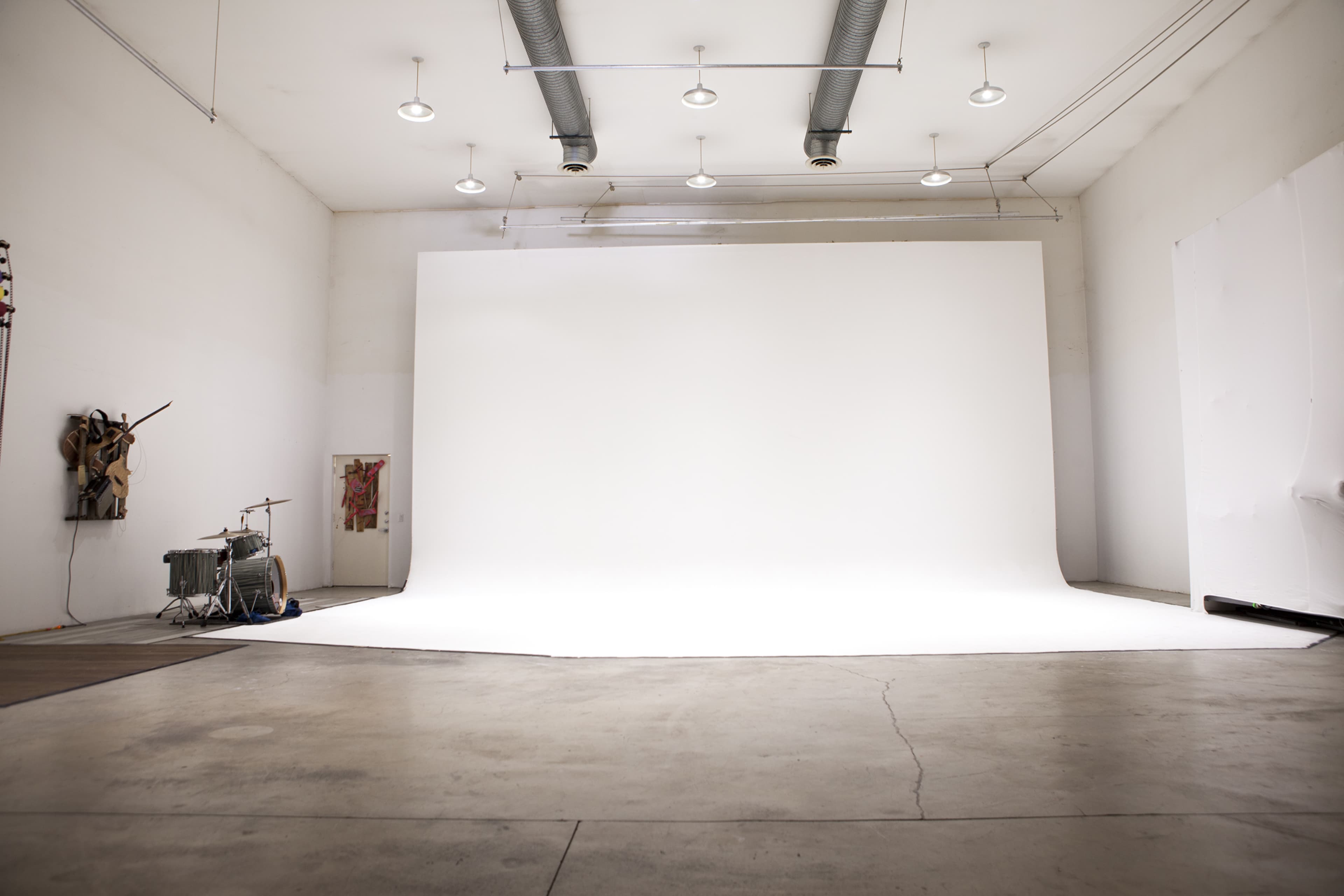 The image shows a large, empty studio space with white walls and a seamless white backdrop, featuring industrial lighting and a drum set in the corner.