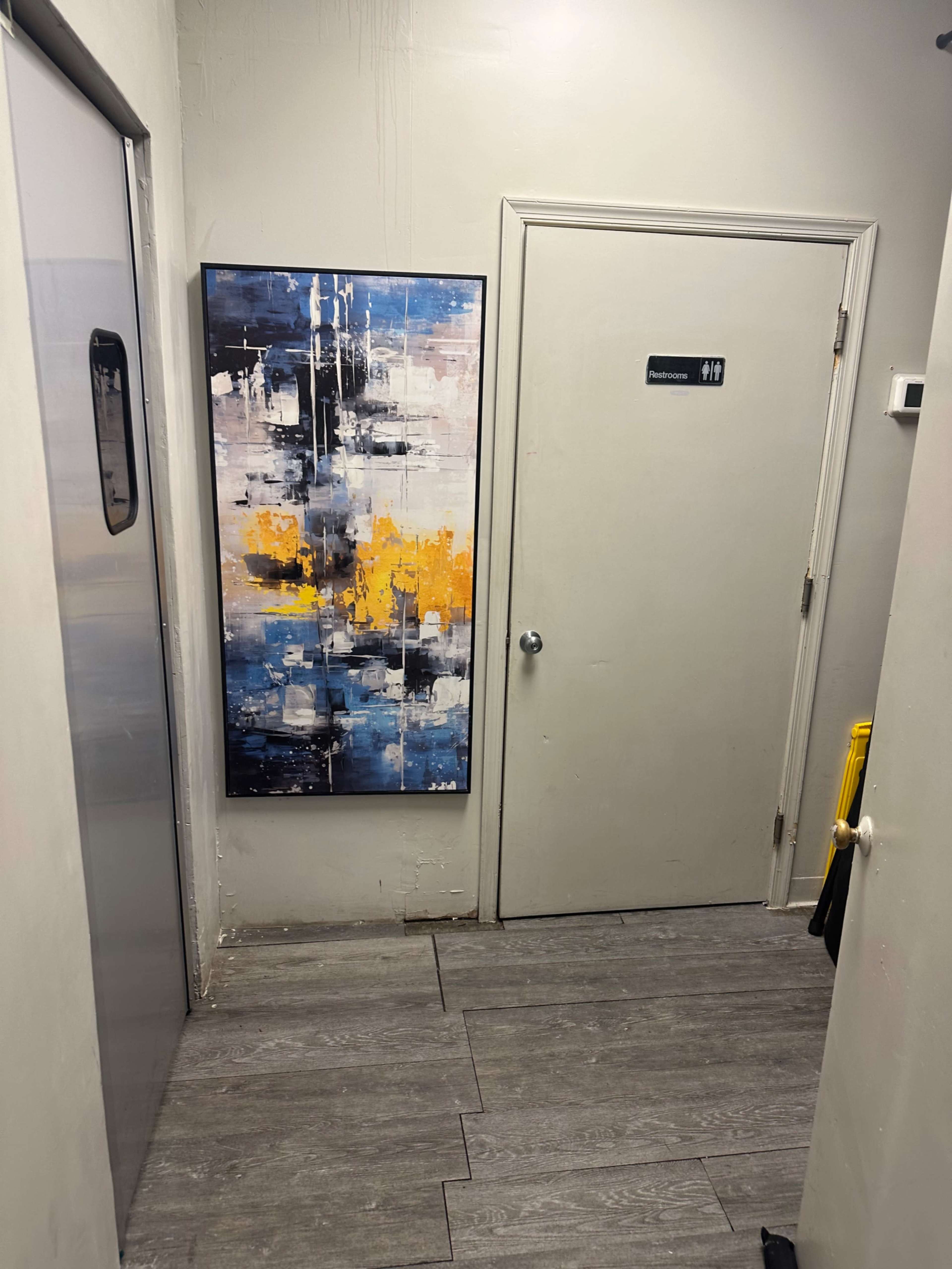The image shows a hallway with a large abstract painting on the wall next to a door labeled for restrooms.