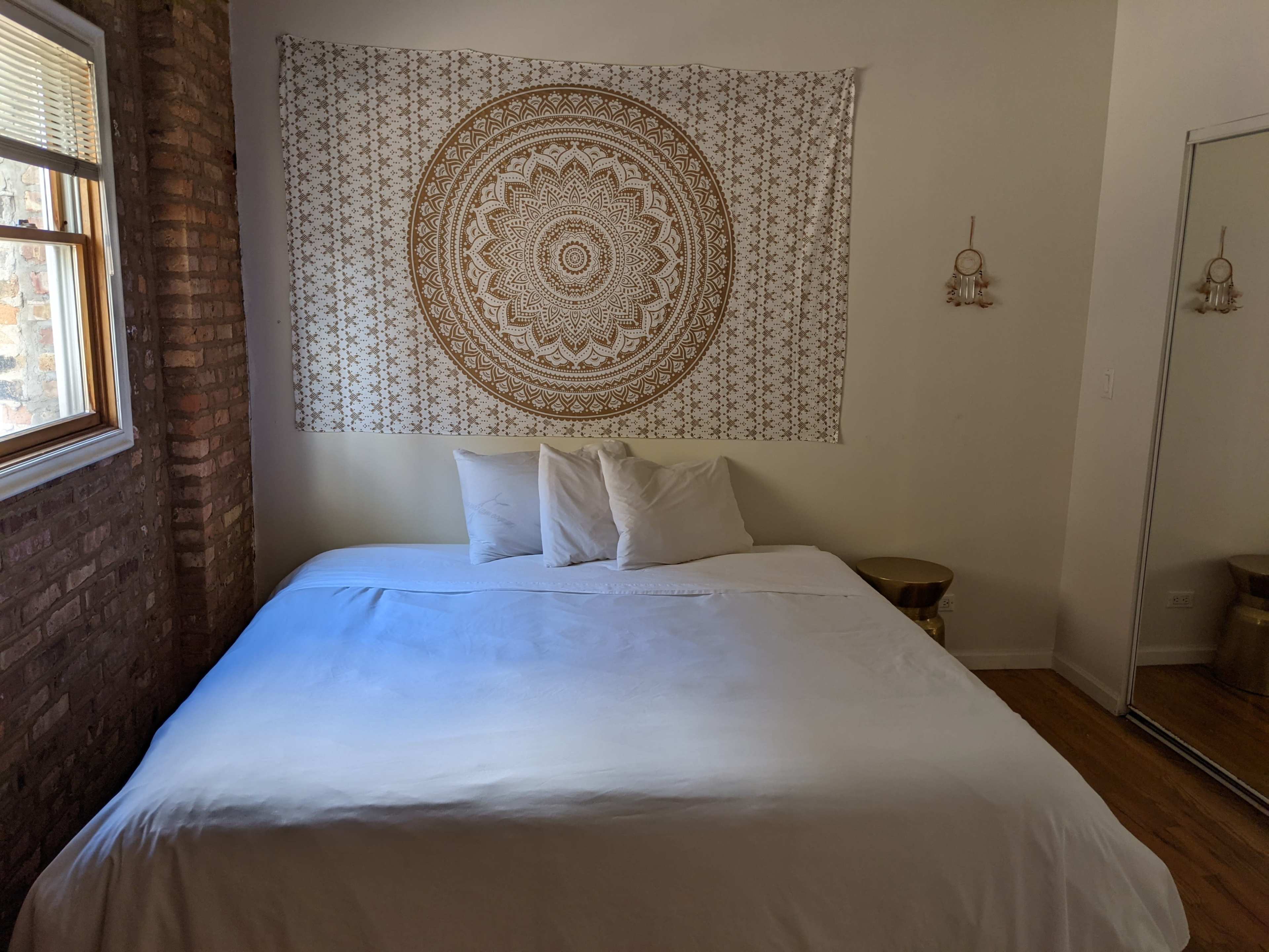 The image shows a bedroom with a bed featuring white bedding, two pillows, and a patterned tapestry hanging on the wall above it, alongside a mirror and a window letting in natural light.
