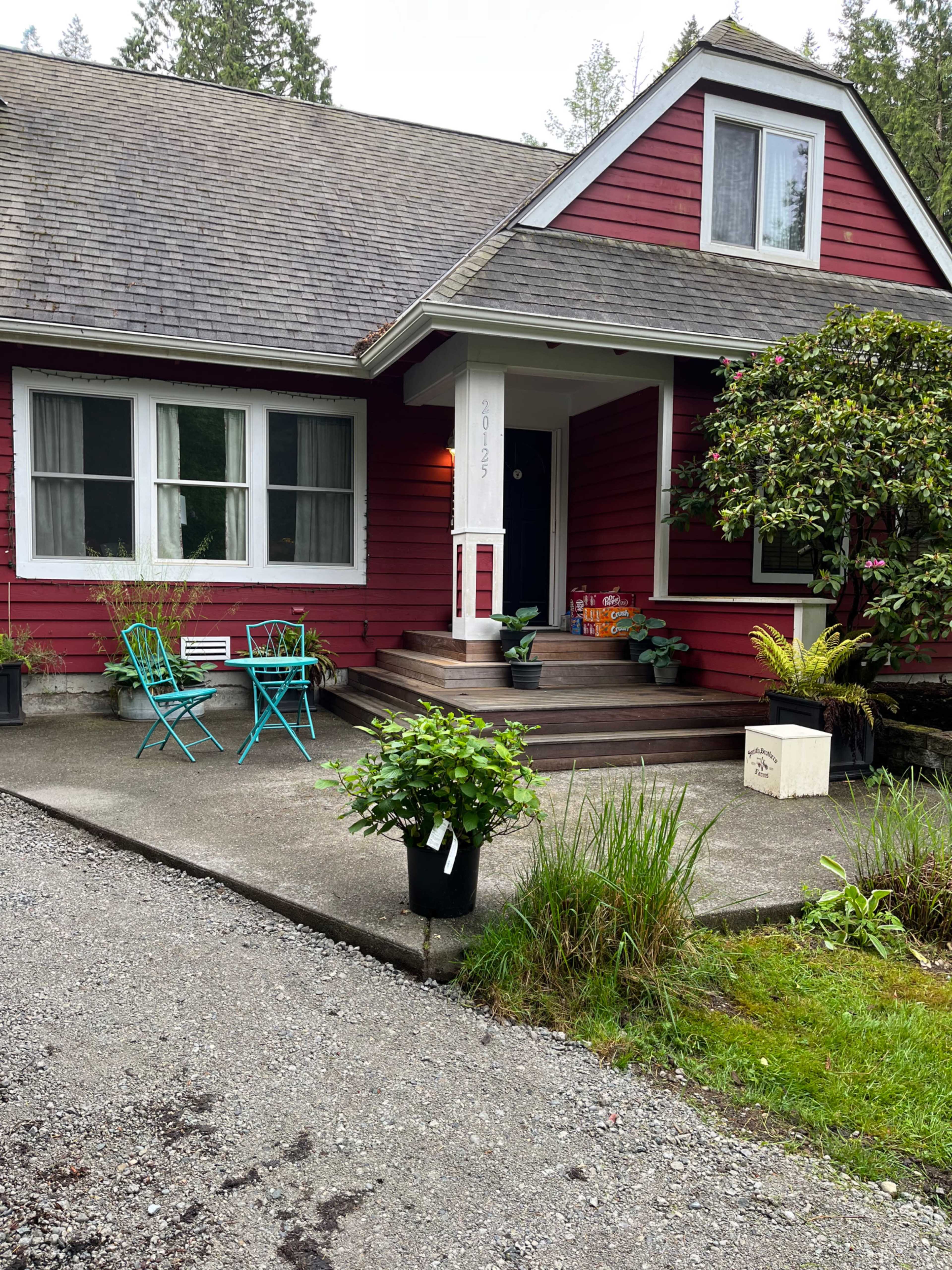 A red house with a front porch features two teal chairs, potted plants, and a gravel driveway lined with greenery.