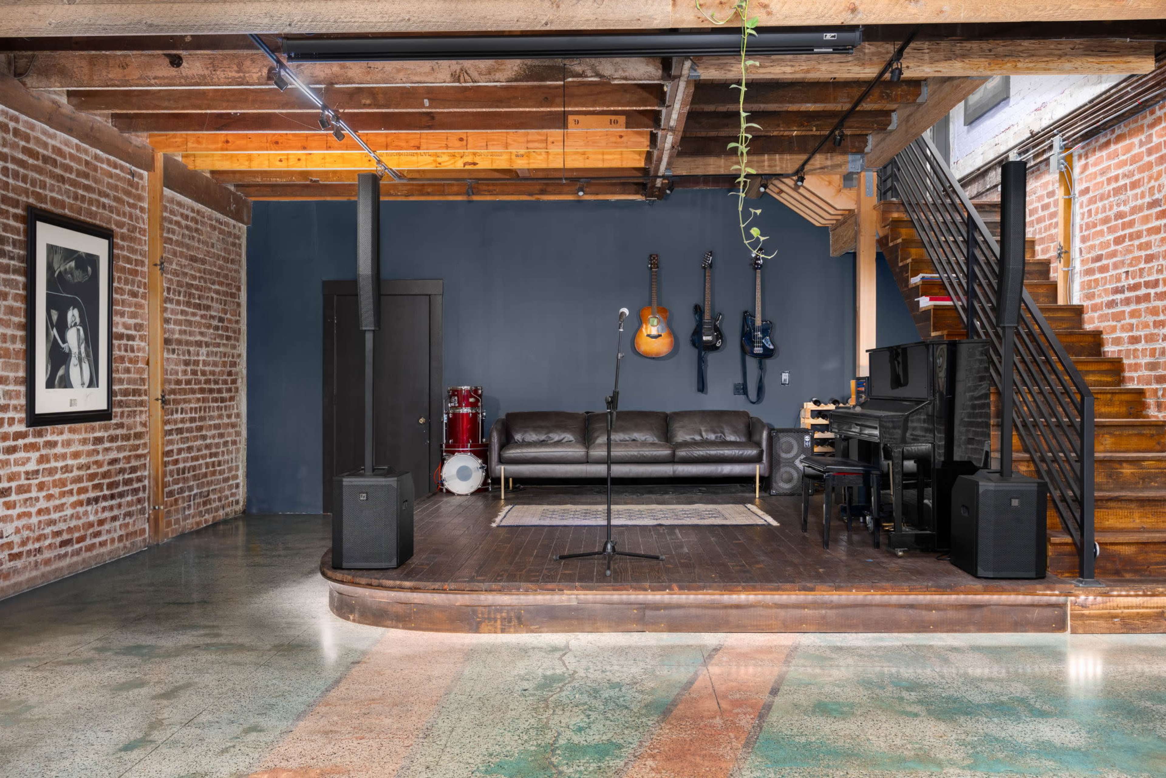 The image shows a spacious music studio with exposed brick walls, a wooden stage area featuring musical instruments, a seating area, and a staircase leading to an upper level.