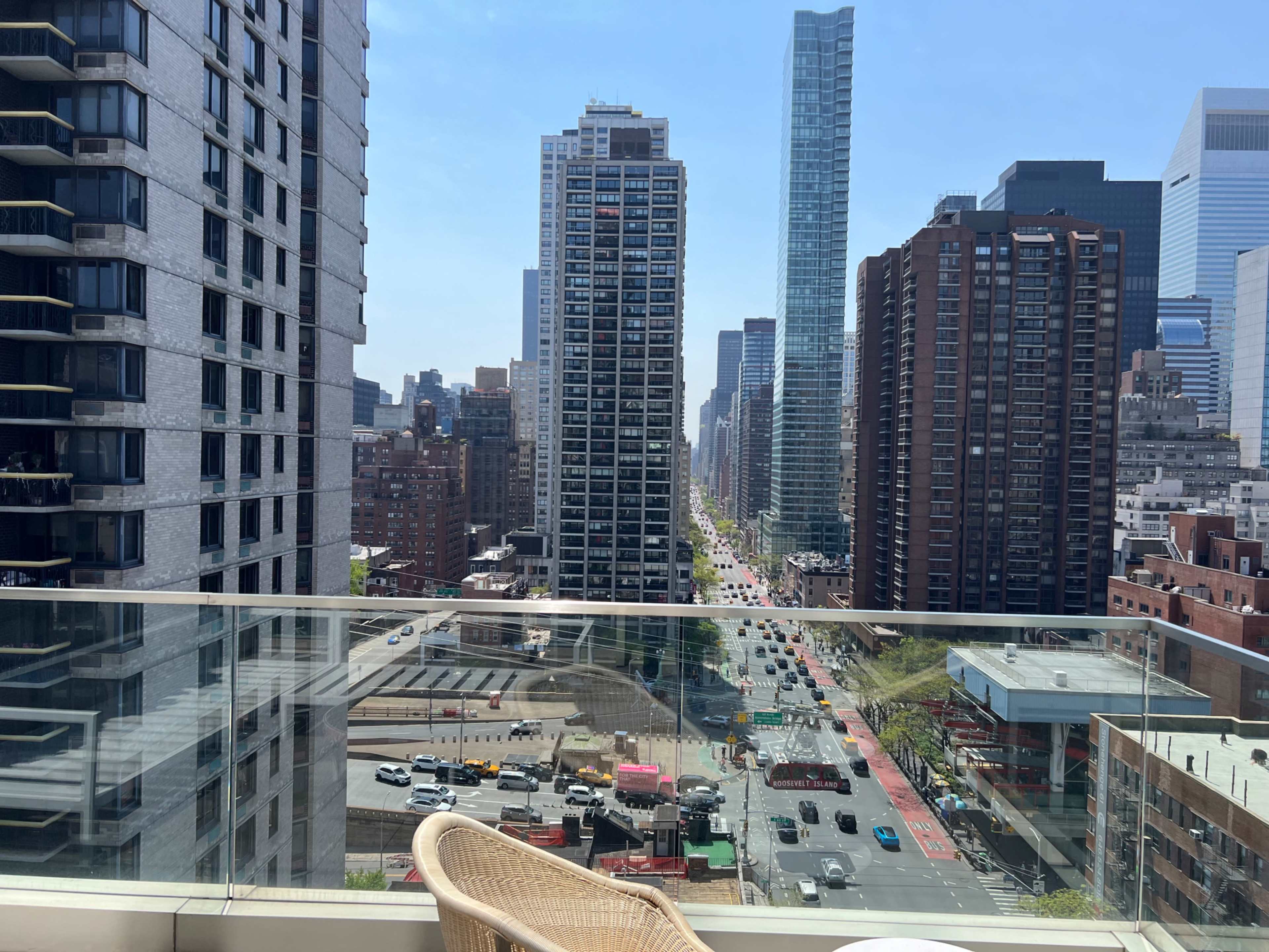 A cityscape with tall buildings lining a busy street, viewed from a balcony with a clear blue sky above.