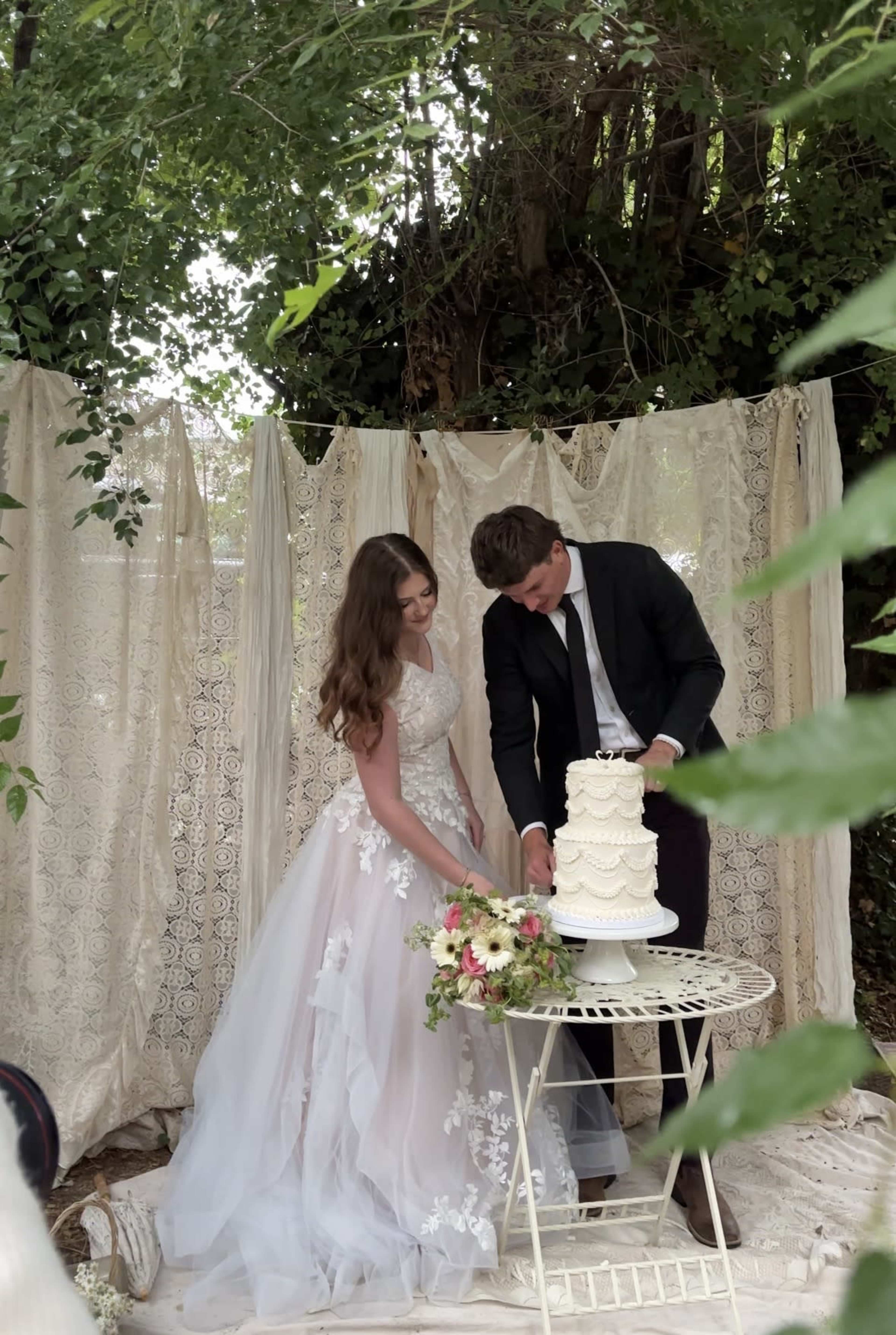 A couple stands together cutting a wedding cake in front of a decorated backdrop with lace curtains and greenery.