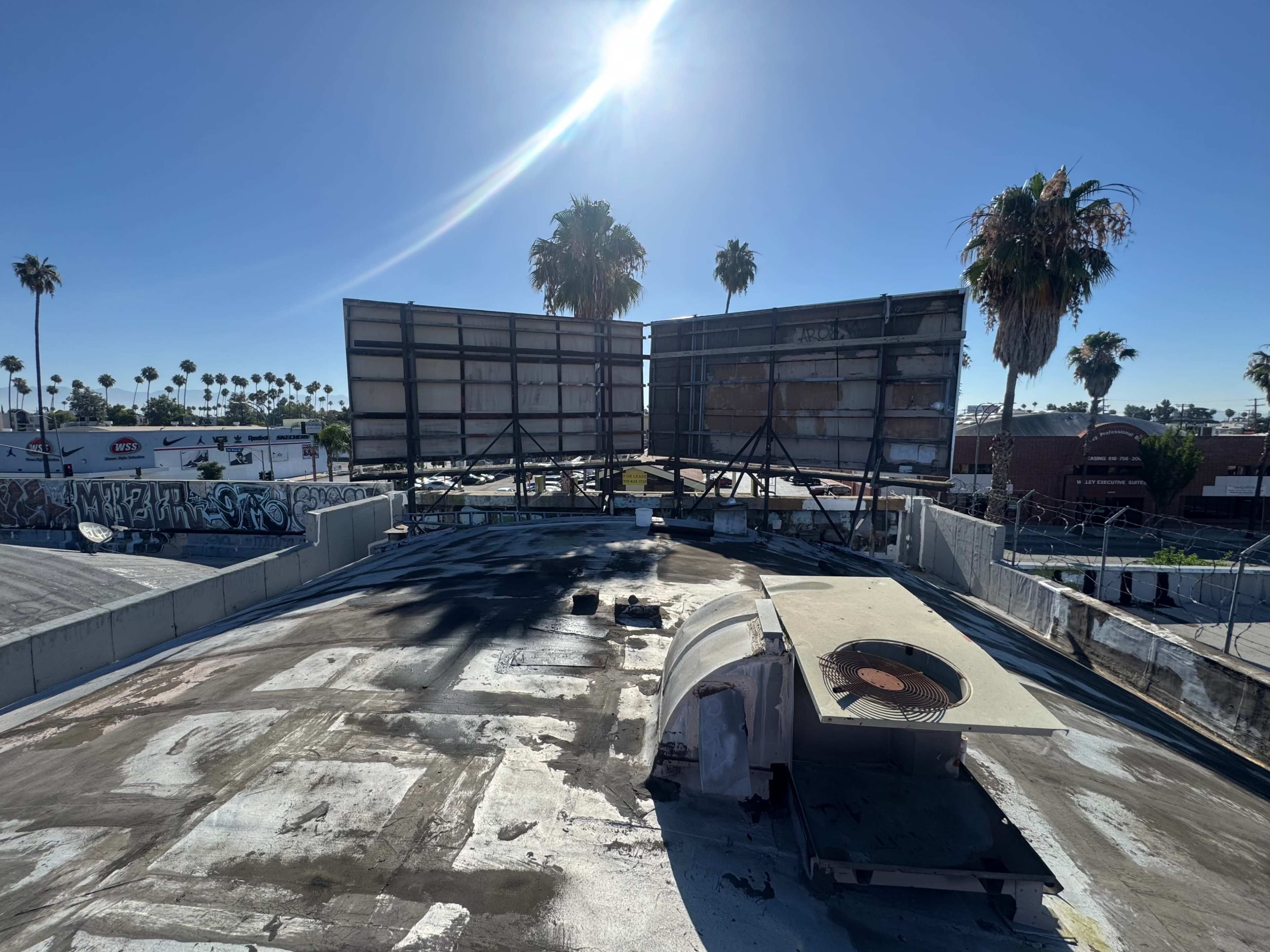 The image shows a rooftop with an air conditioning unit, two large billboards in the background, and palm trees lining the skyline under a bright sun.