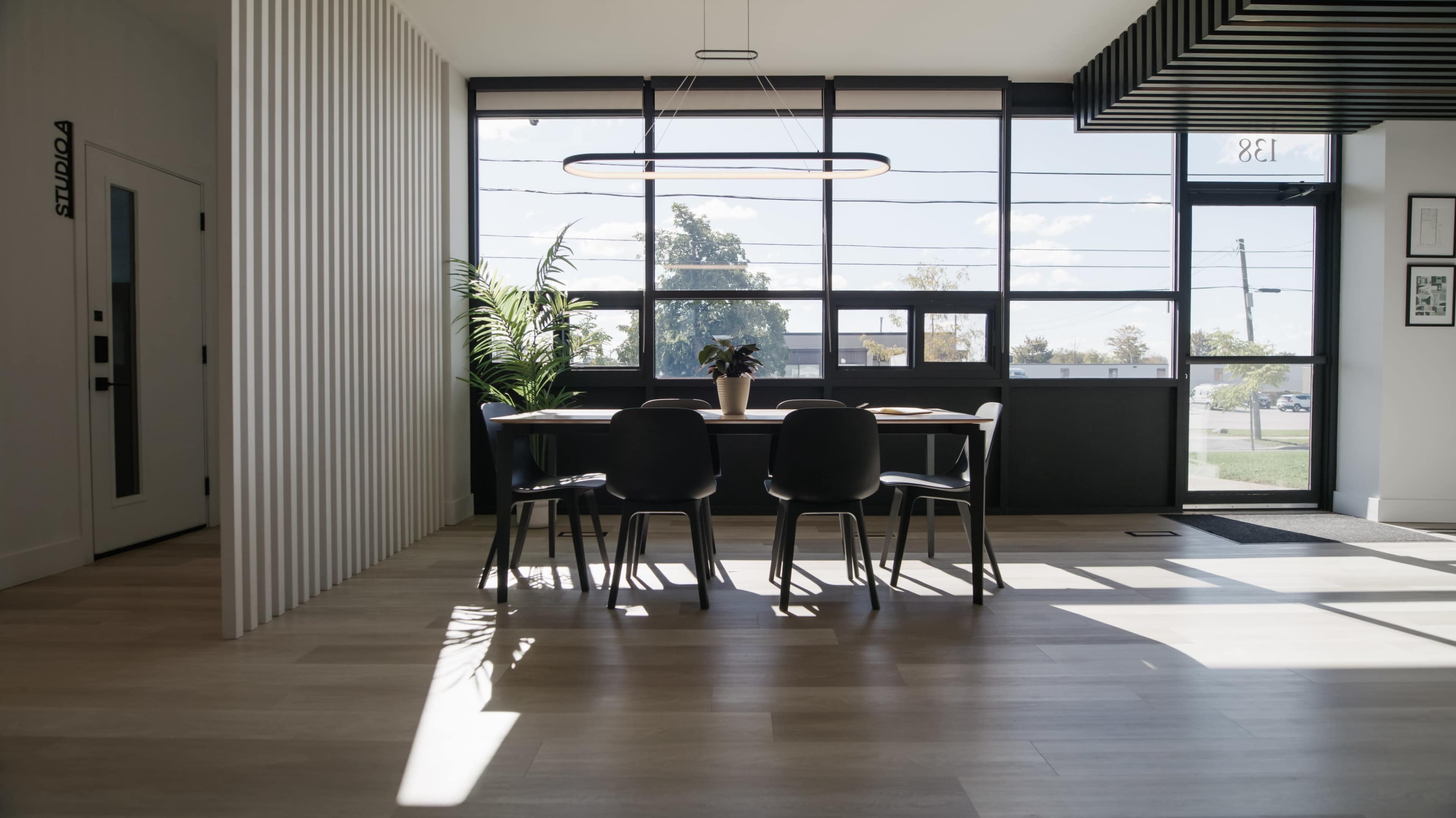 A dining area features a large table surrounded by black chairs, illuminated by natural light from large windows.