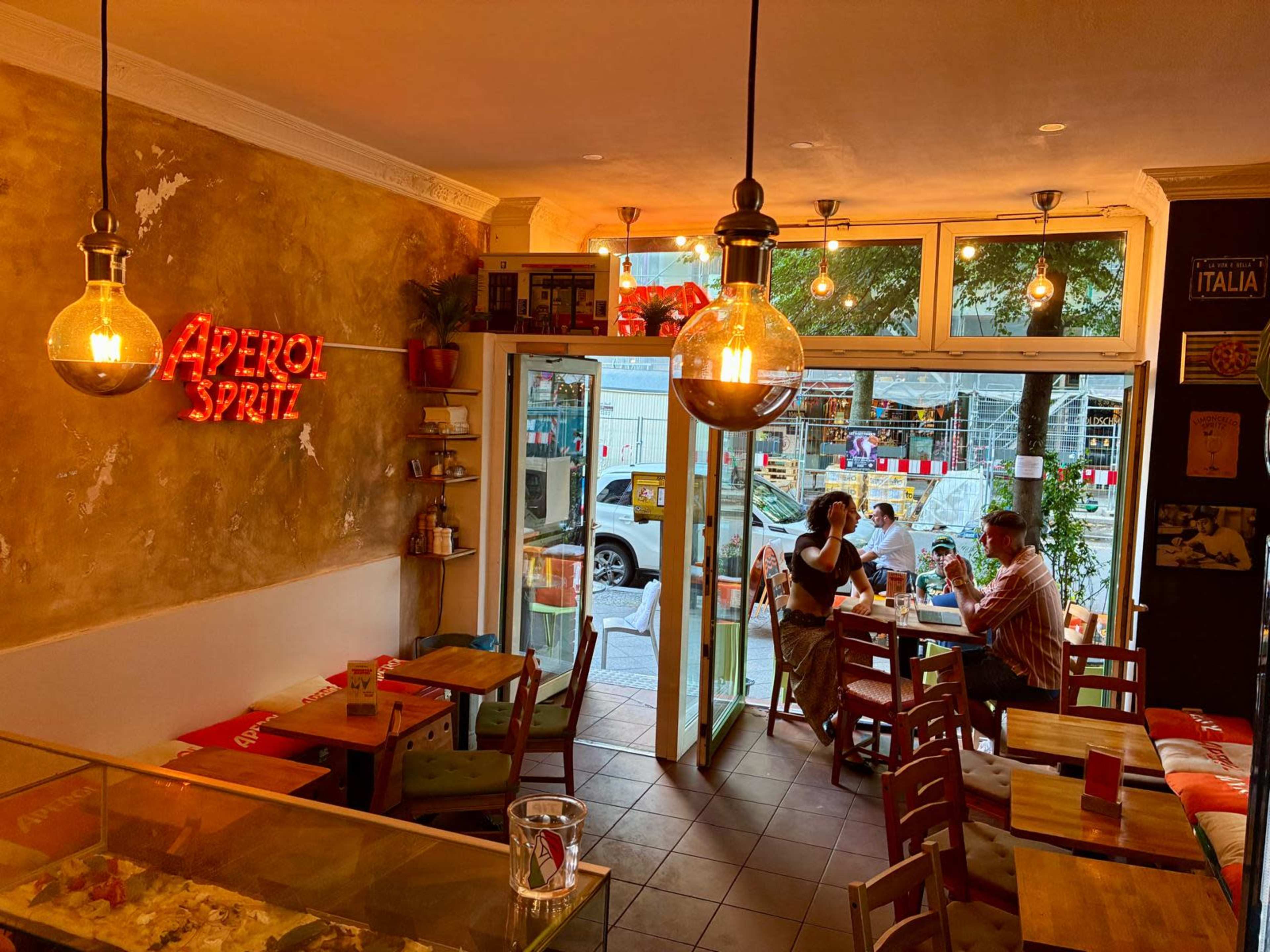 The image shows a cozy café interior with a retro light fixture, a neon sign for Aperol Spritz, and two people sitting at a table while a door opens to the outside street.