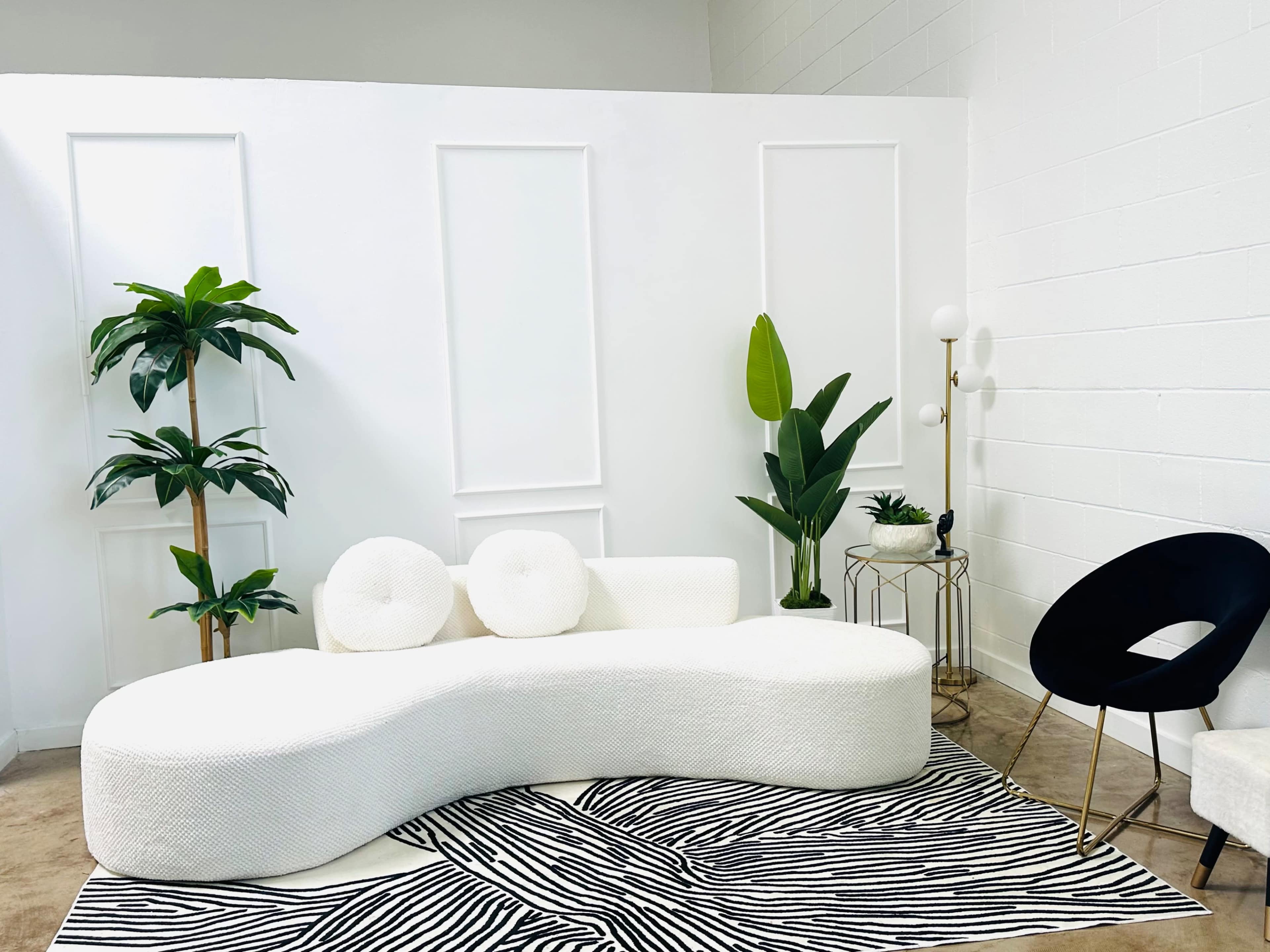 The scene features a modern, white curved sofa set against a white wall, accompanied by various indoor plants and a patterned rug.