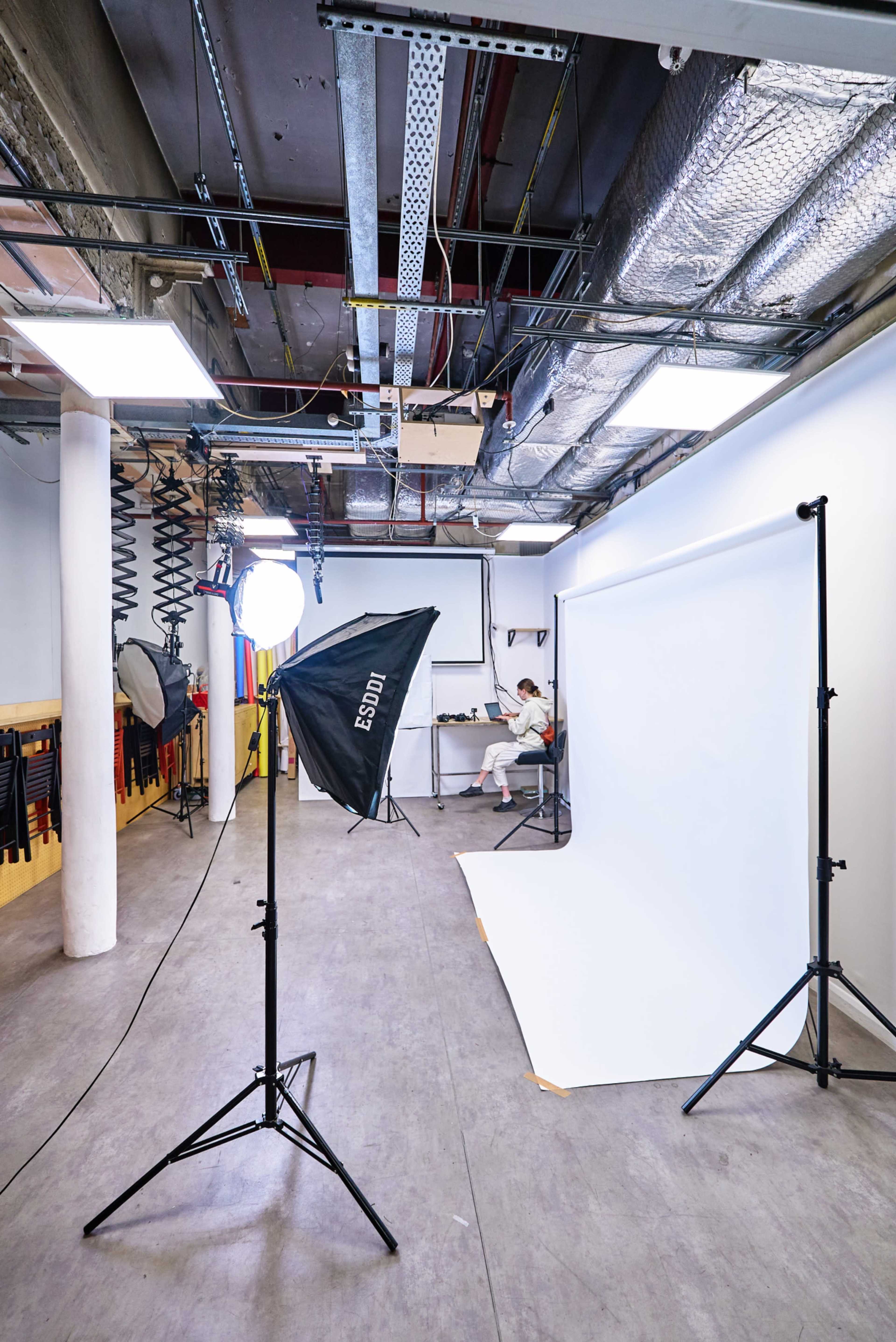 The image shows a photography studio featuring a seamless background setup, lighting equipment, and a person seated at a desk.