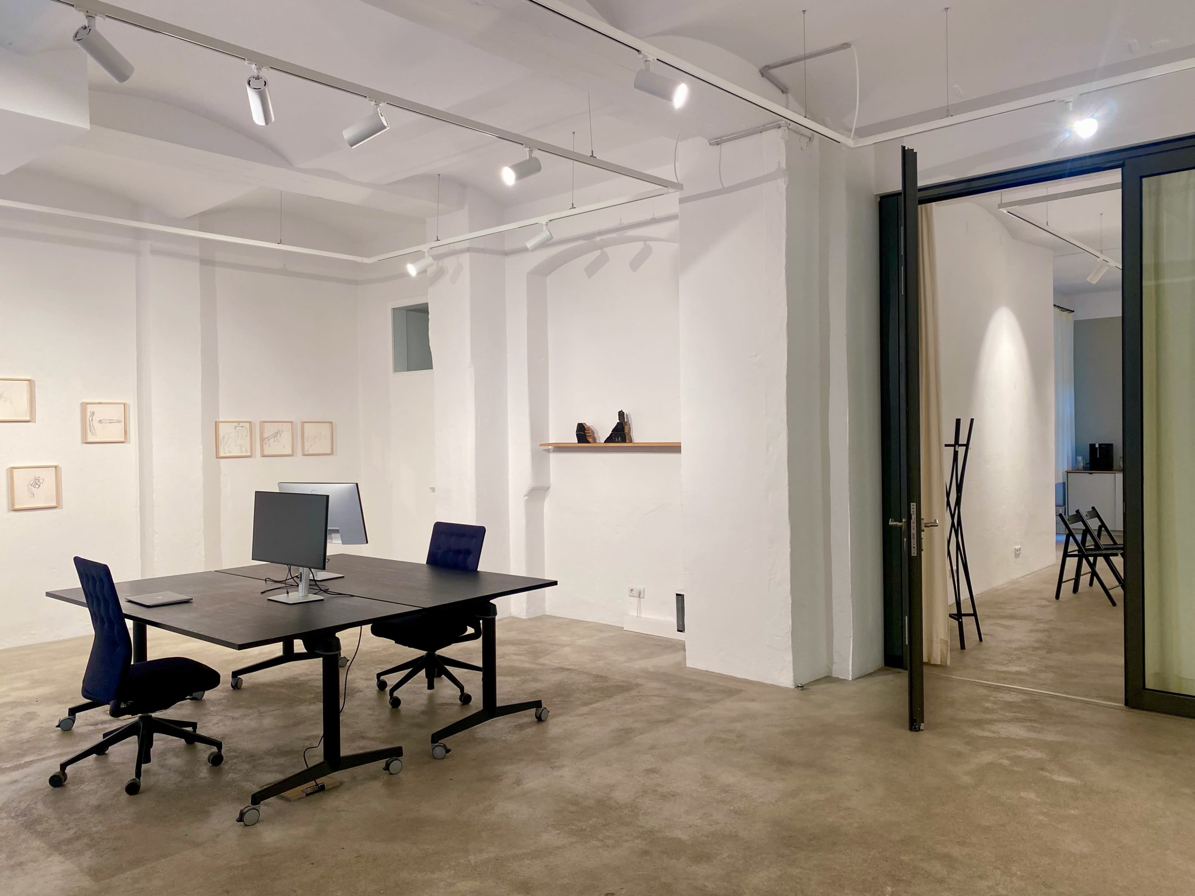 A minimalist office space features a dark table with a computer and two blue chairs, surrounded by white walls and minimal artwork.