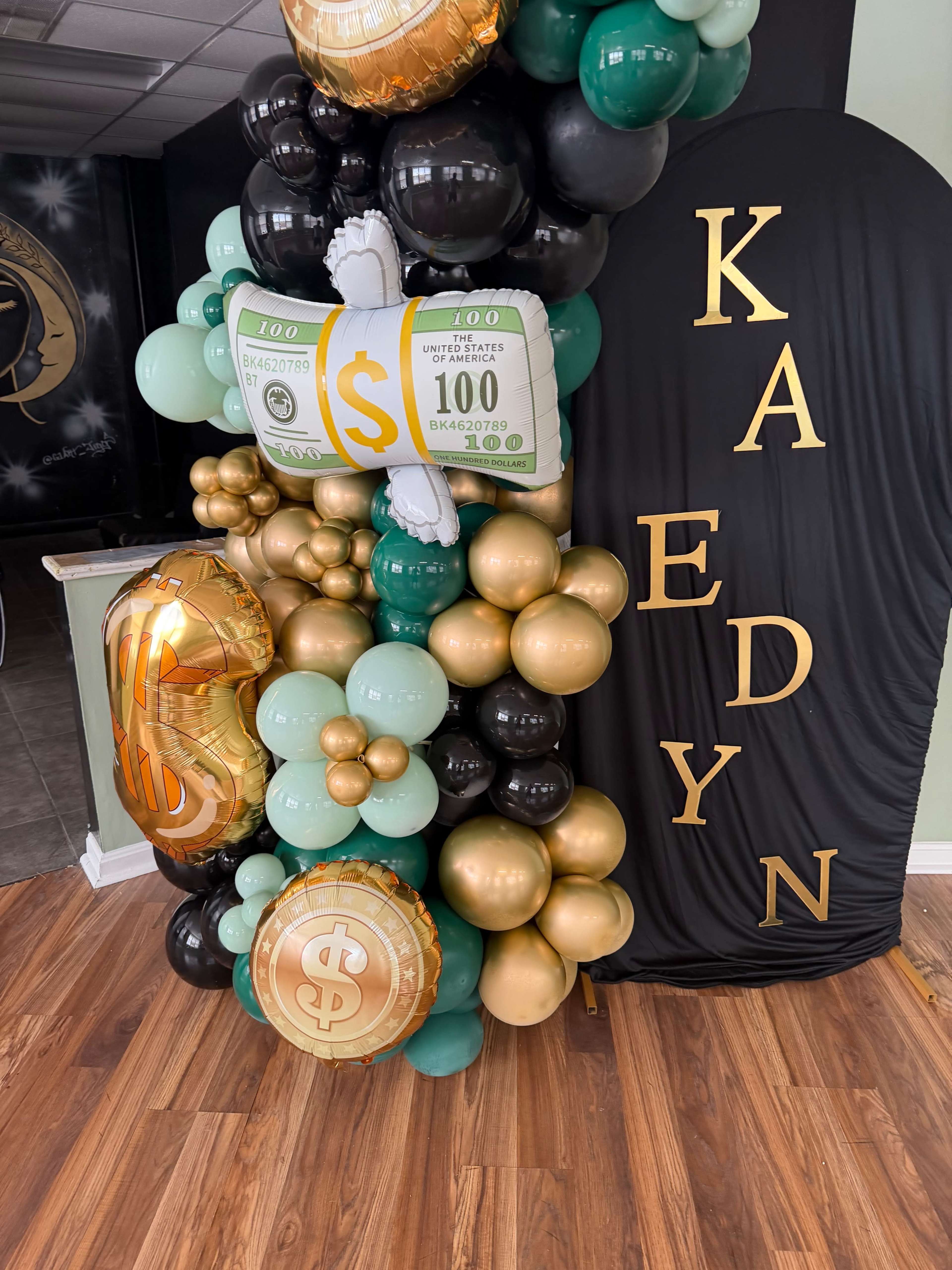 The image features a vibrant balloon arrangement in shades of green, gold, black, and bronze, with a dollar sign motif and a decorative stand displaying the name "KADYN."