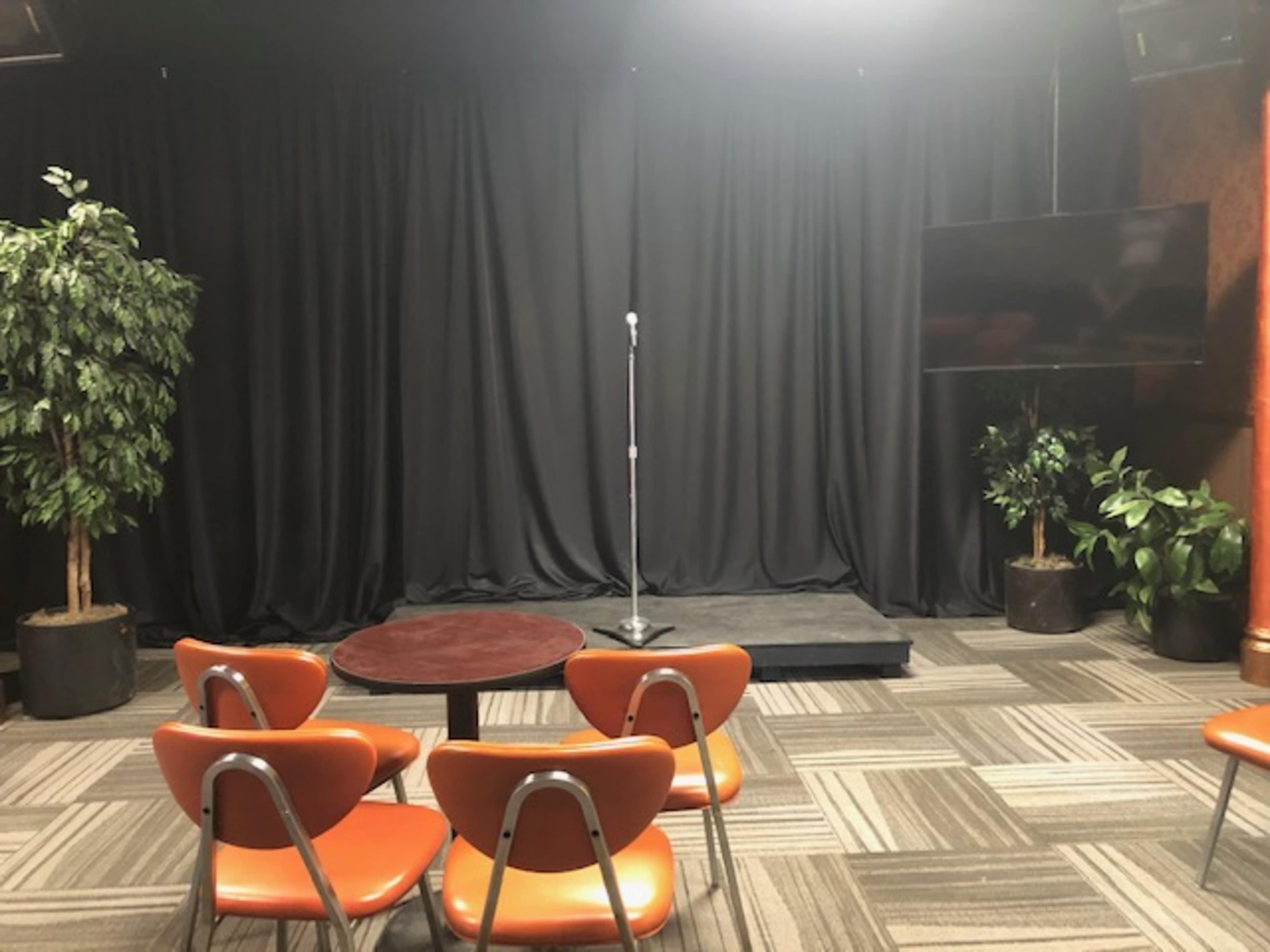 A small performance area features a microphone on a stand against a black curtain, with orange chairs arranged around a round table.