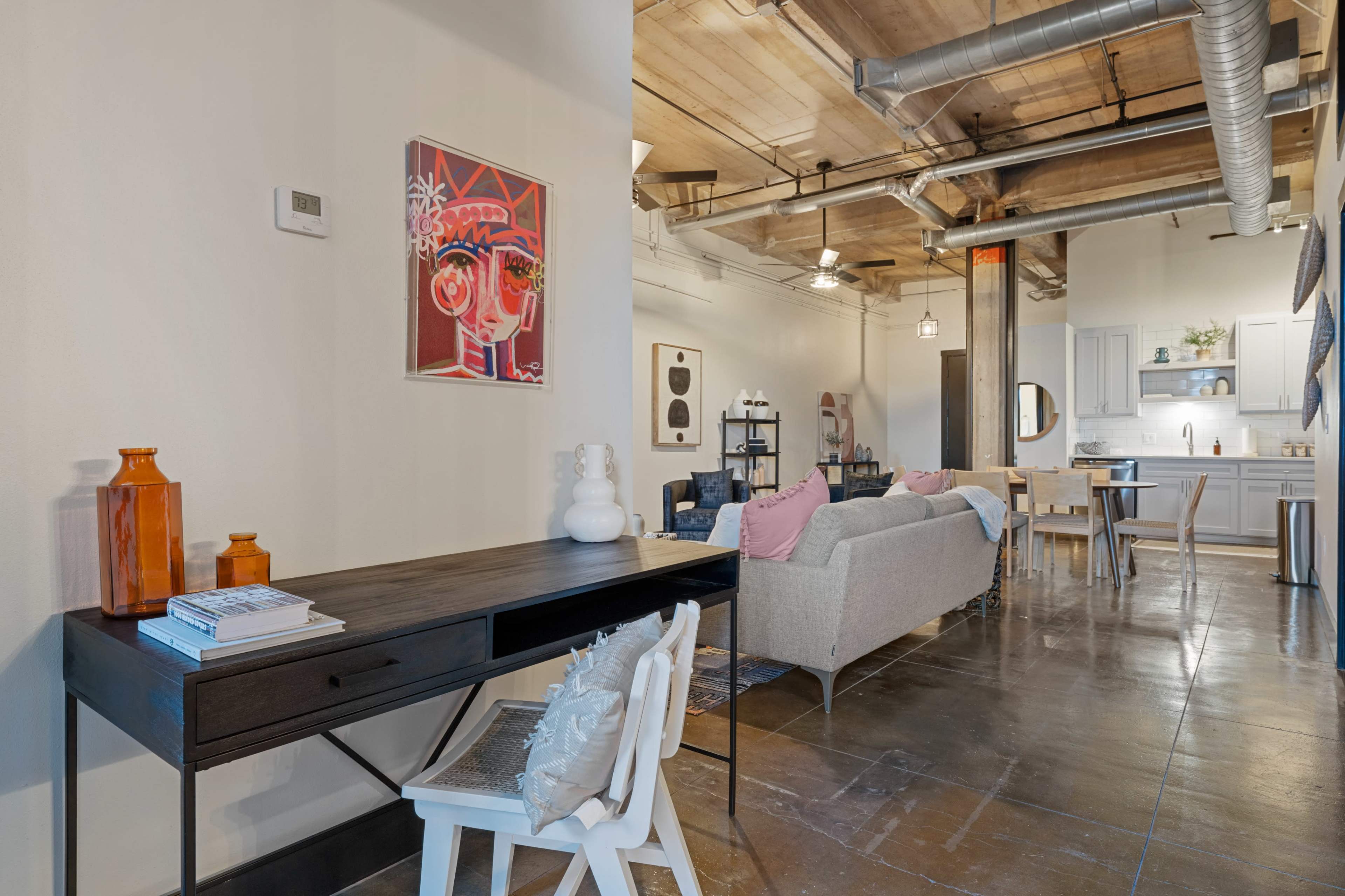 A modern interior space featuring a desk and chair in the foreground, with a sofa and dining area visible in the background.