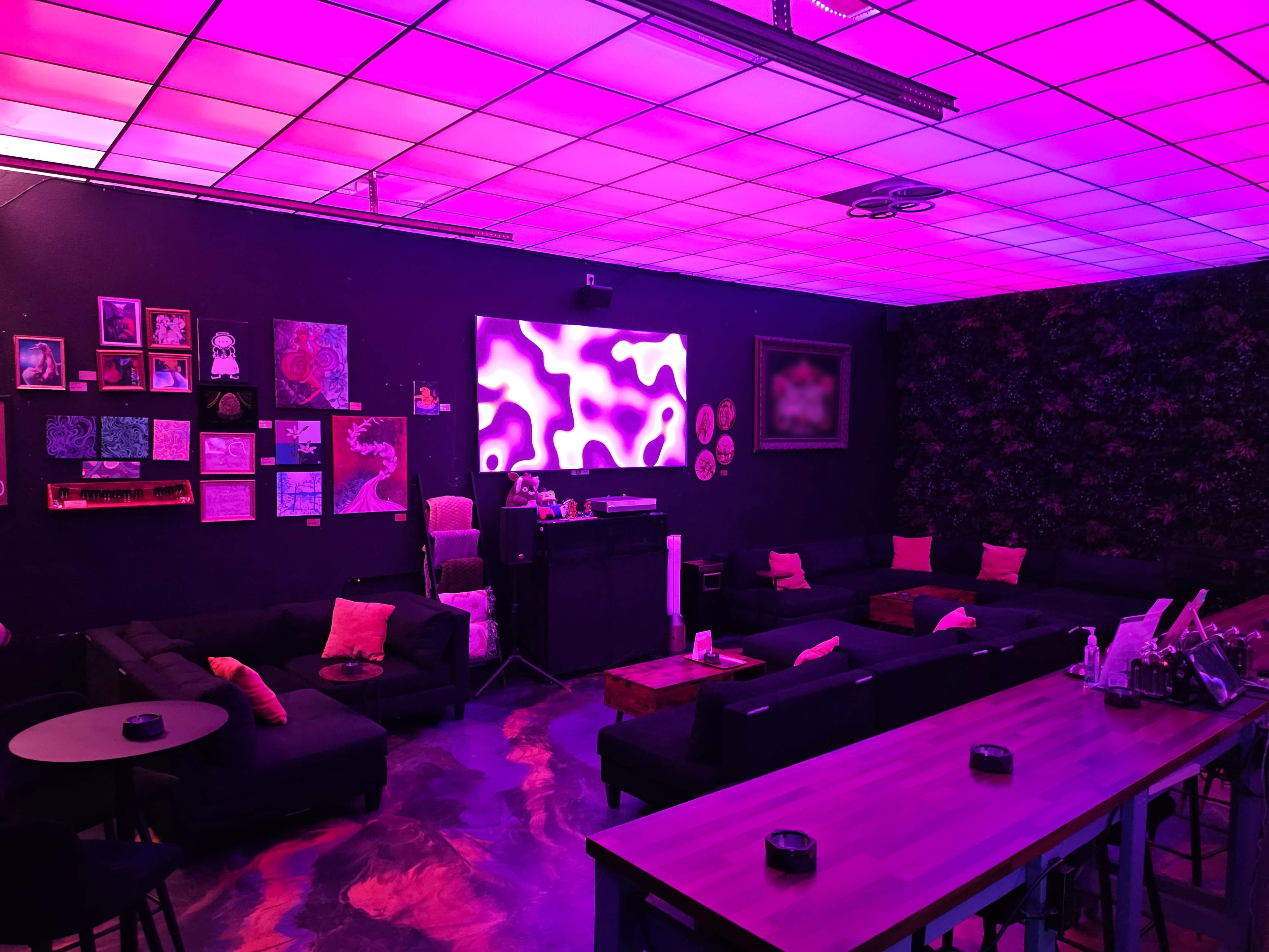 The image shows a dimly lit lounge with purple lighting, featuring black sofas, a long wooden table, and colorful artwork displayed on the walls.