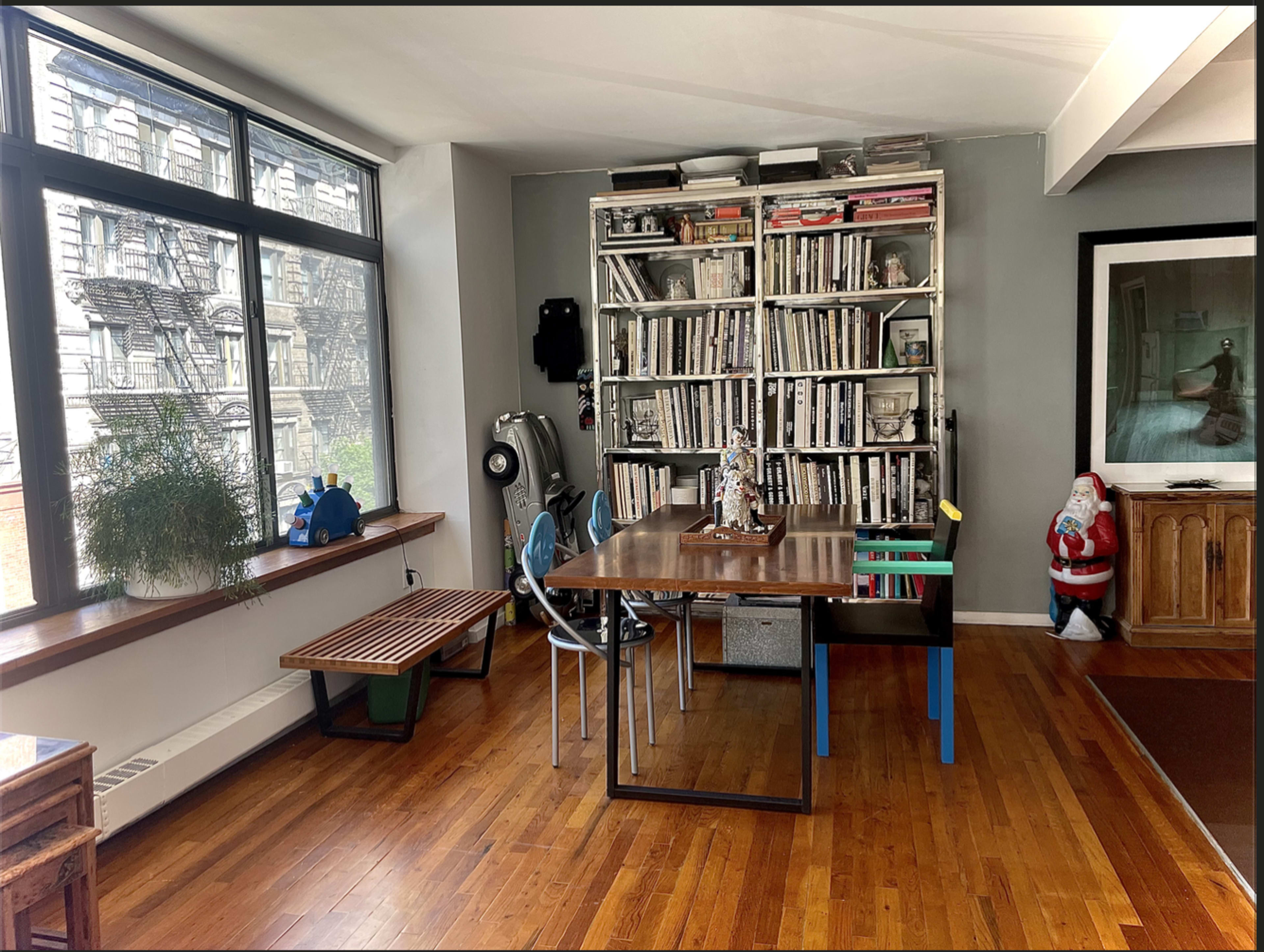 The image shows a modern interior space with a wooden dining table, chairs of various colors, a large bookshelf filled with books, and a Santa decoration in the corner.