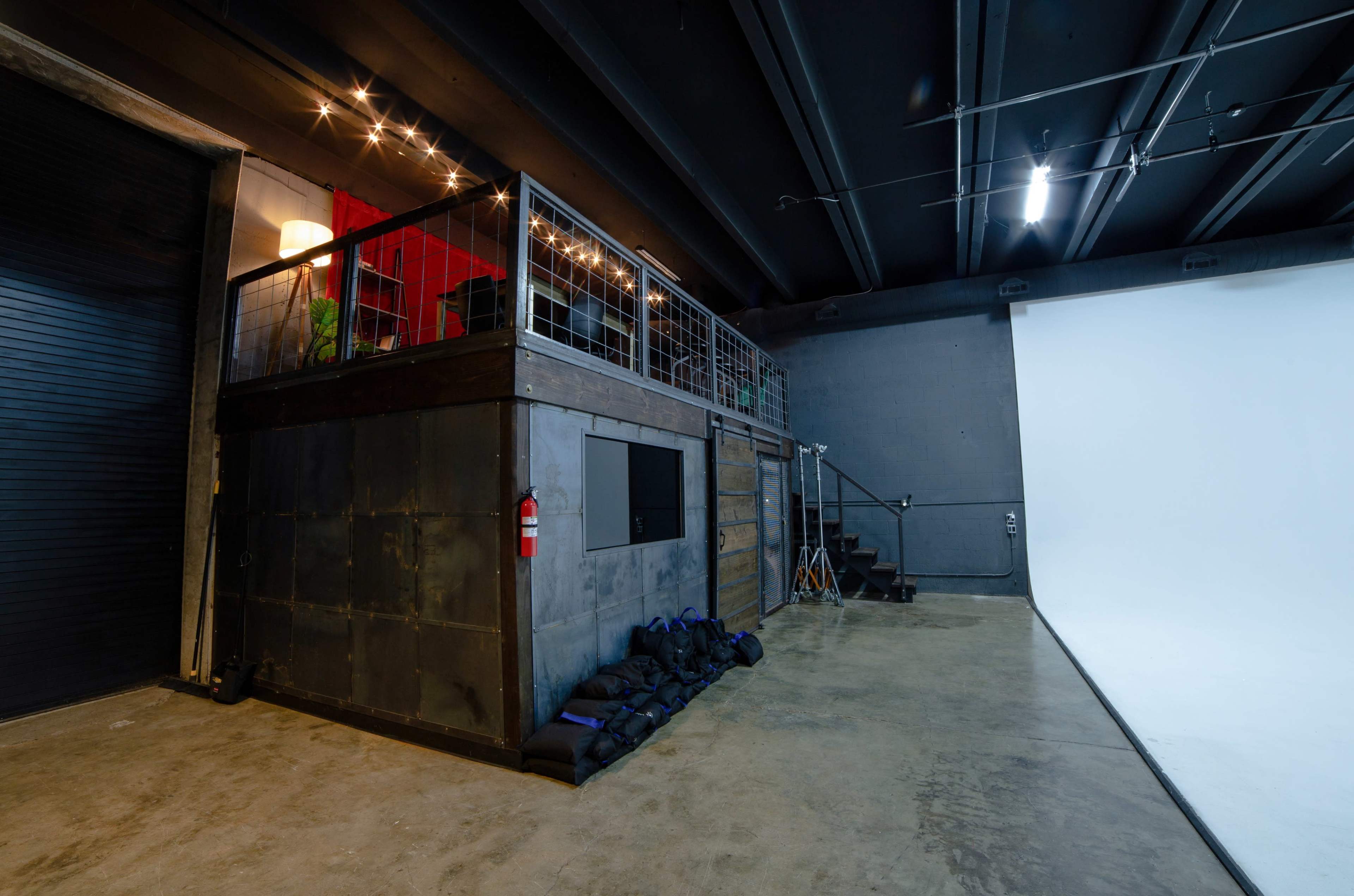 The image shows a spacious studio with a multi-level area featuring a metal and wood structure, a staircase, and a blank white backdrop on one side.
