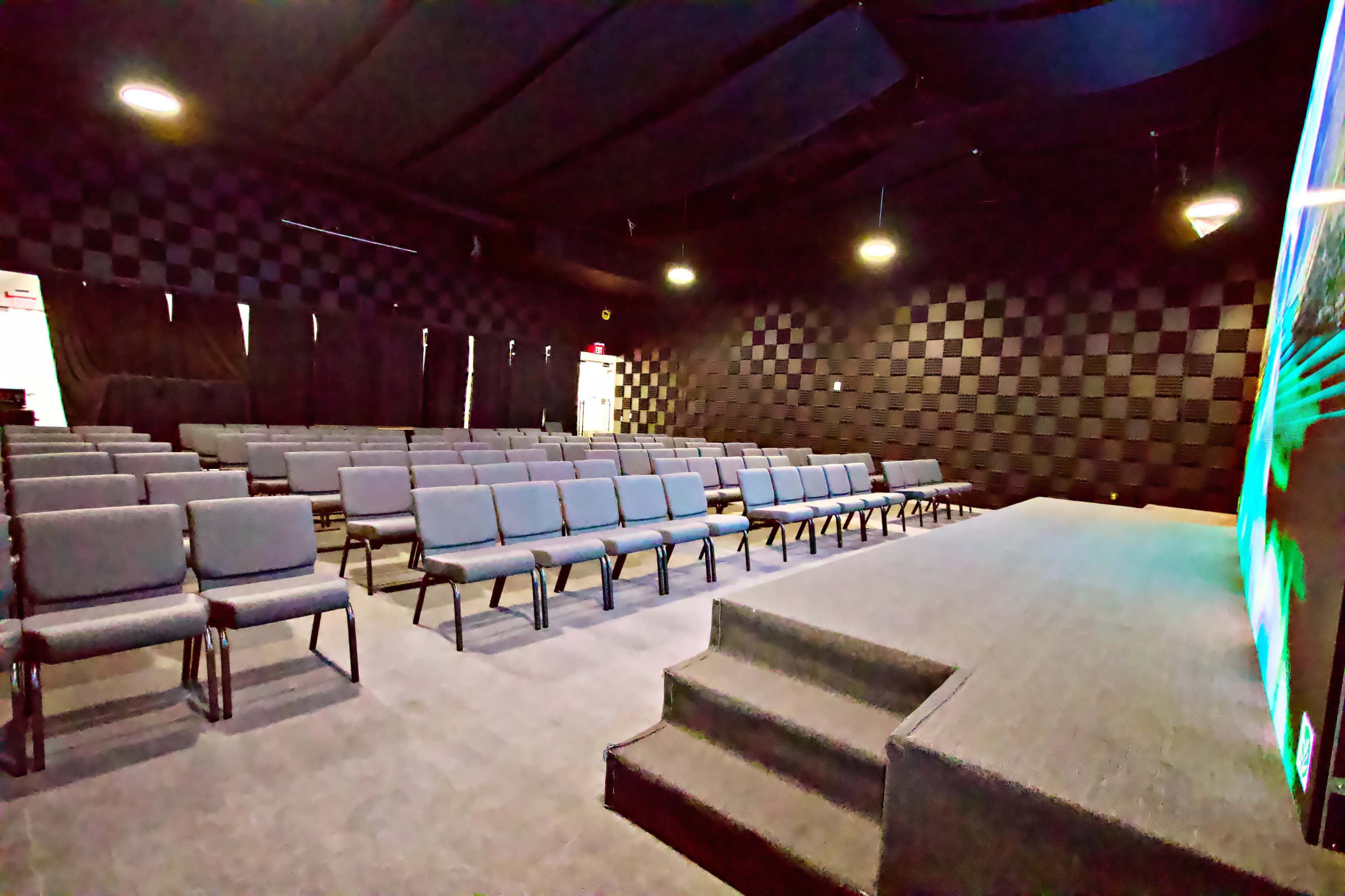 An empty auditorium with rows of gray chairs facing a small stage, all set against a black and gray checkered wall.