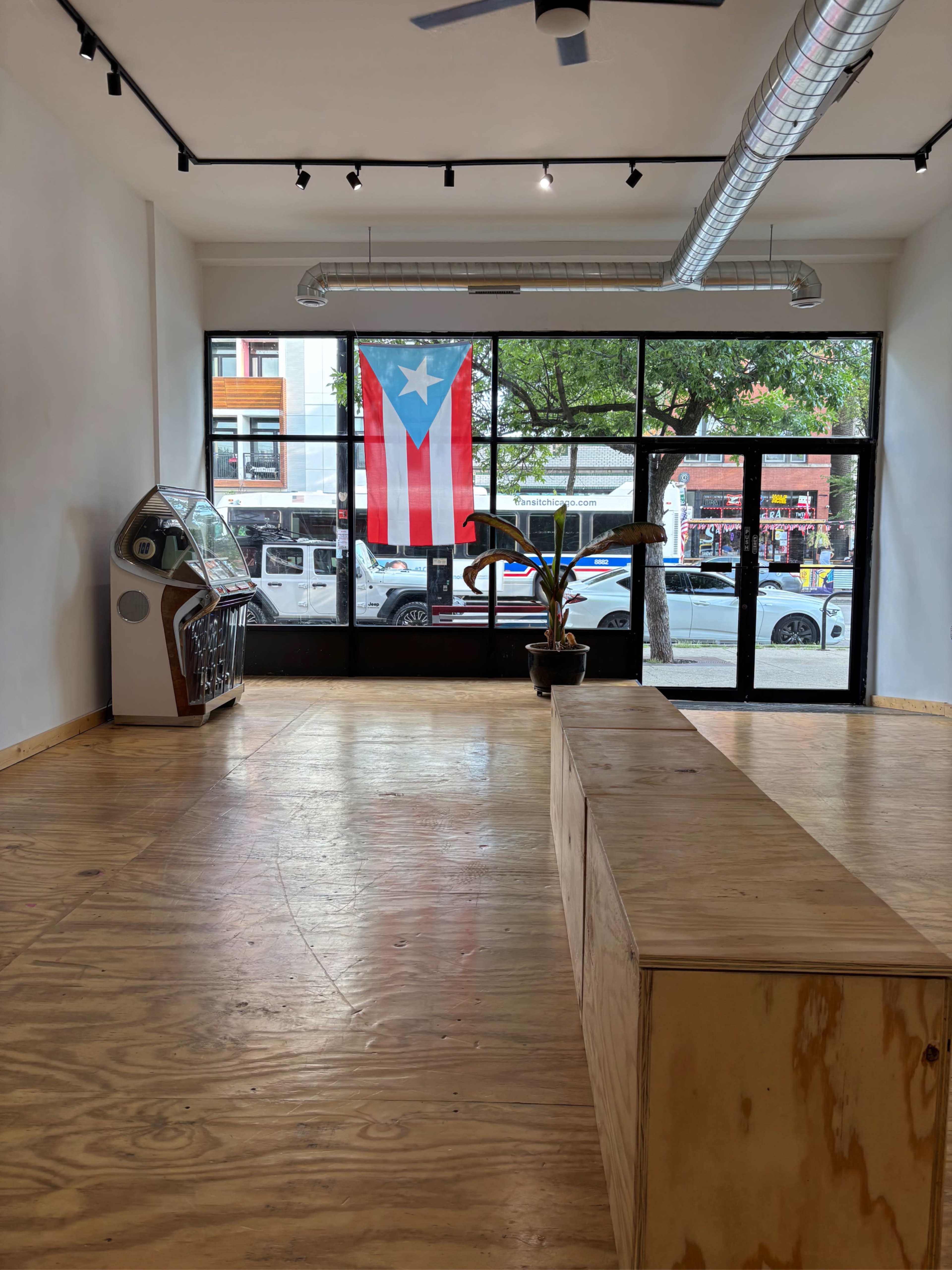 The image shows an empty retail space featuring a wooden platform, a vintage arcade machine in the corner, and a Puerto Rican flag displayed in the window.