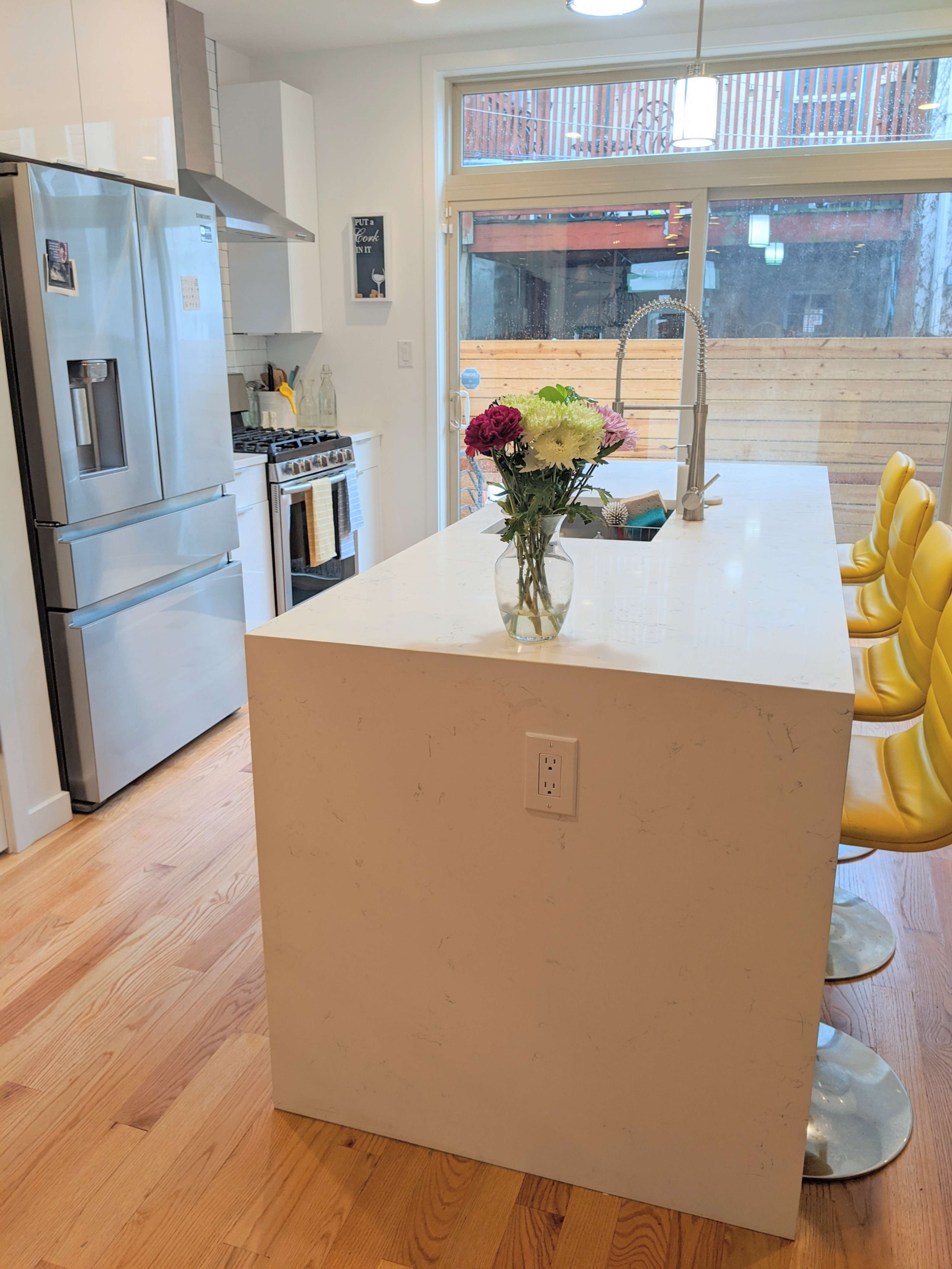 A modern kitchen features stainless steel appliances, a large white island with a vase of flowers, and yellow bar stools.