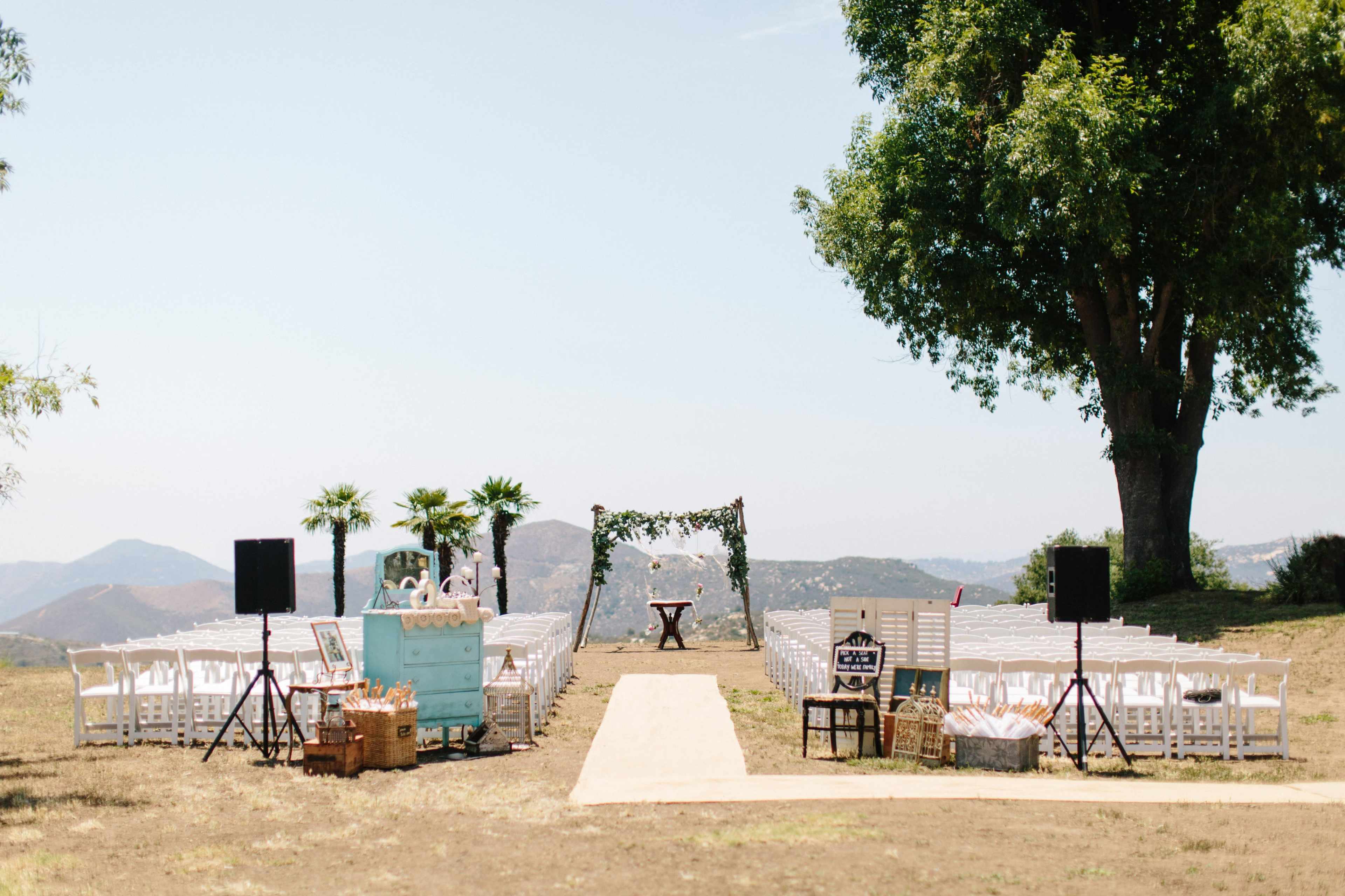 An outdoor wedding ceremony setup features white chairs arranged in rows along a central aisle leading to an arch adorned with greenery and flowers, set against a mountainous backdrop.