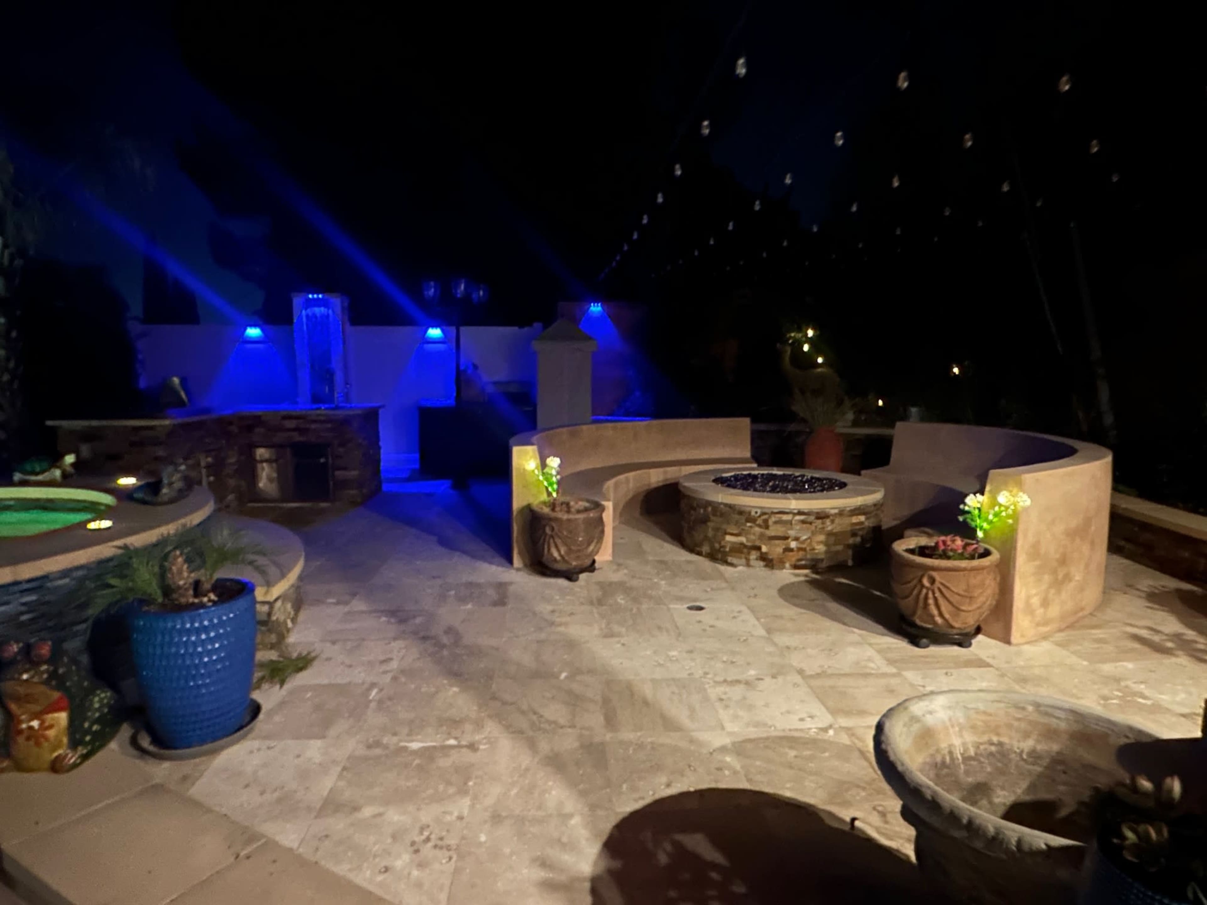 The image shows a well-lit outdoor patio featuring curved seating, a stone fire pit, and decorative plants under string lights at night.