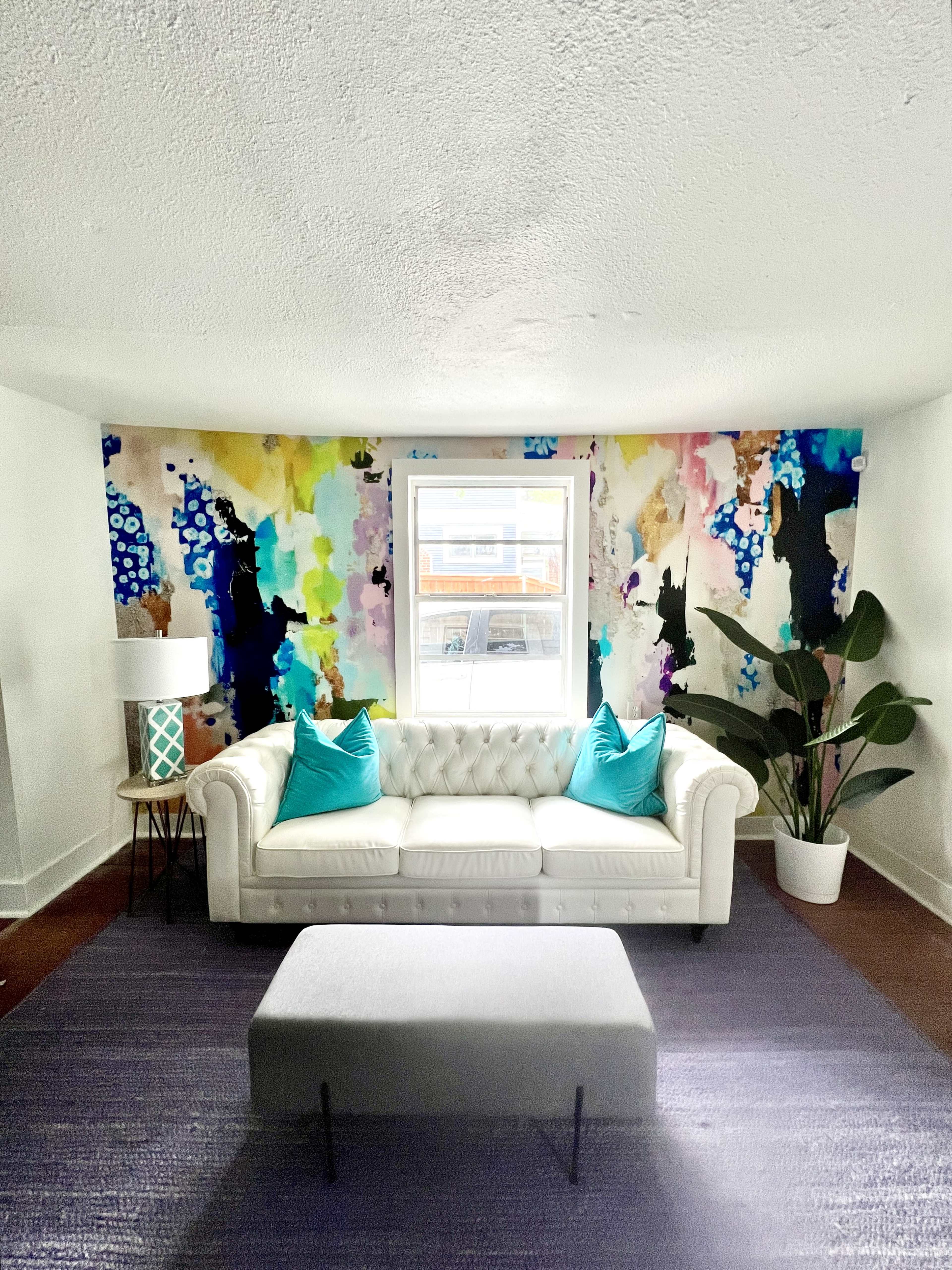 A white tufted sofa with turquoise cushions is positioned facing a window, surrounded by colorful abstract wall art and a potted plant.