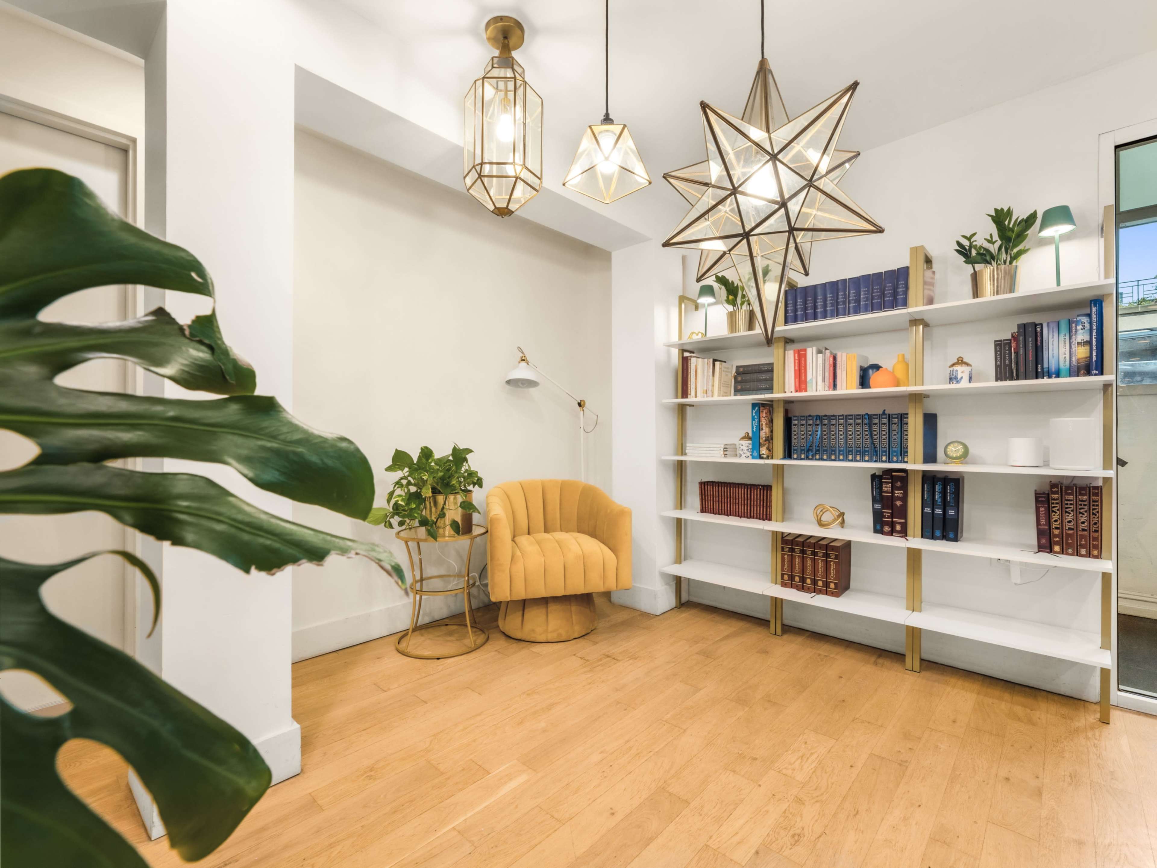 The image features a modern, well-lit interior with a yellow armchair, a white bookshelf filled with books, and decorative plants, all accented by geometric pendant lights.