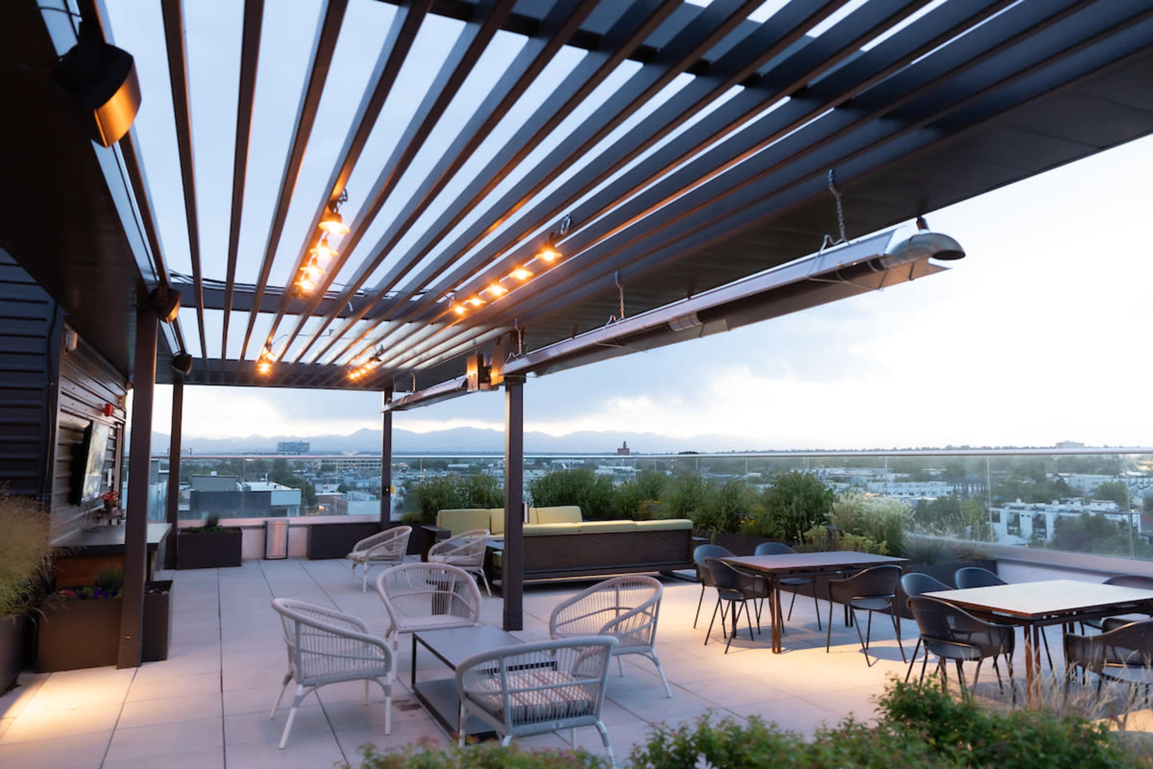 The image shows a rooftop terrace featuring modern furniture, overhead lighting, and a view of the surrounding landscape.