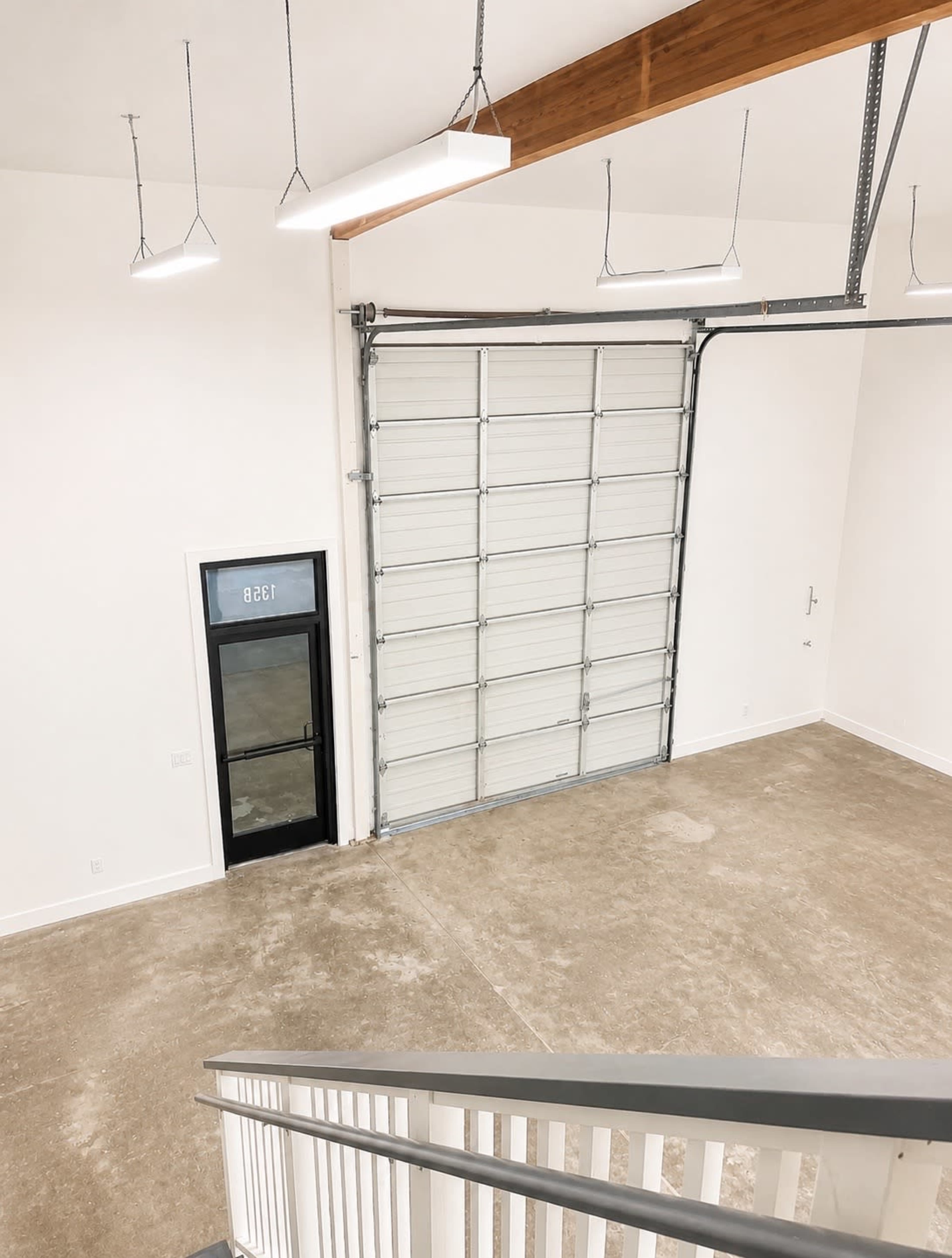 The image shows a spacious, empty room with a large garage door and a glass entry door, featuring a staircase leading down to the concrete floor.