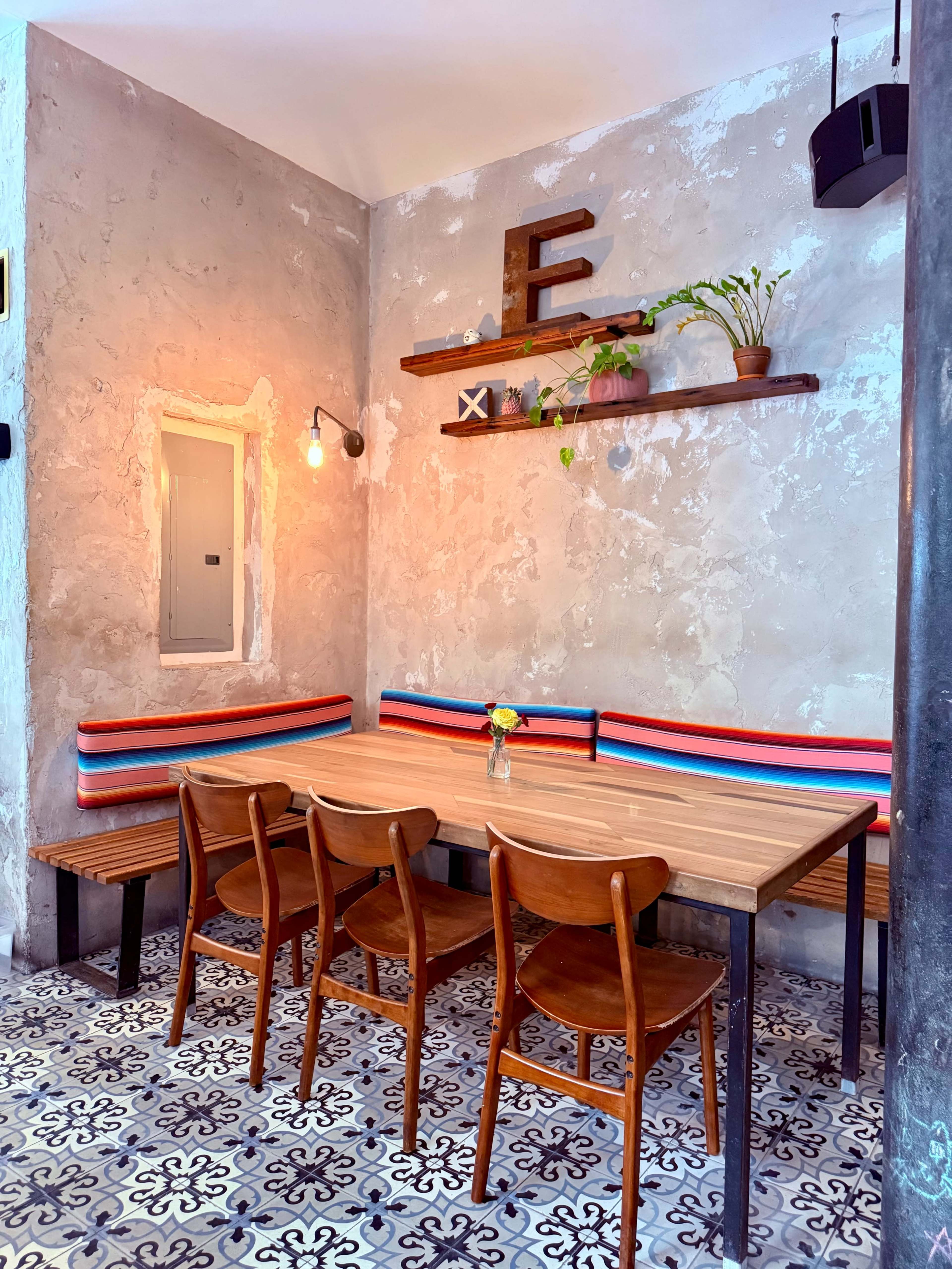 A wooden table surrounded by four chairs is placed against a textured wall with shelves holding plants and decorative items in a café setting.