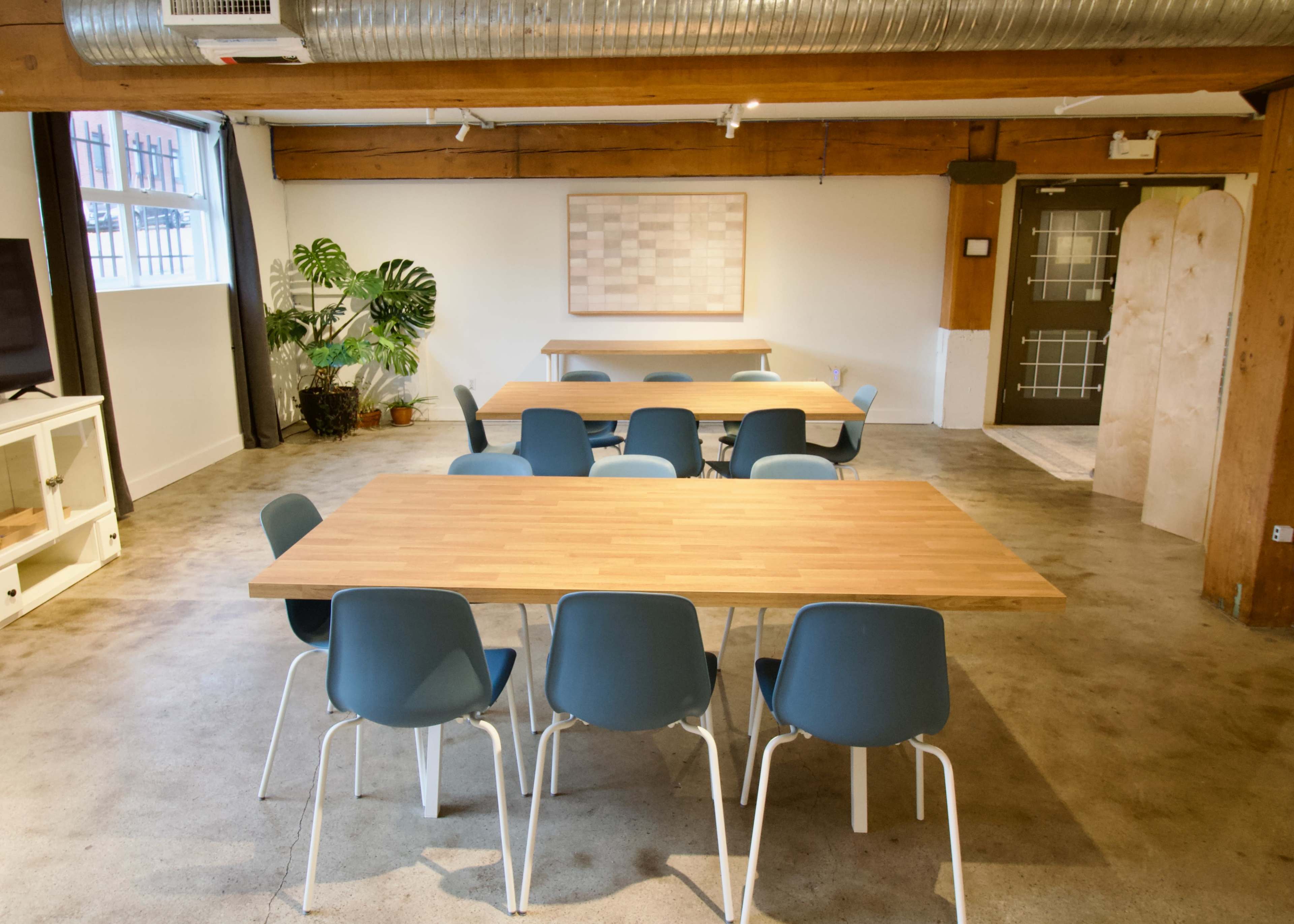 The image shows a modern workspace featuring two wooden tables surrounded by blue chairs, with a large window and a plant in the background.