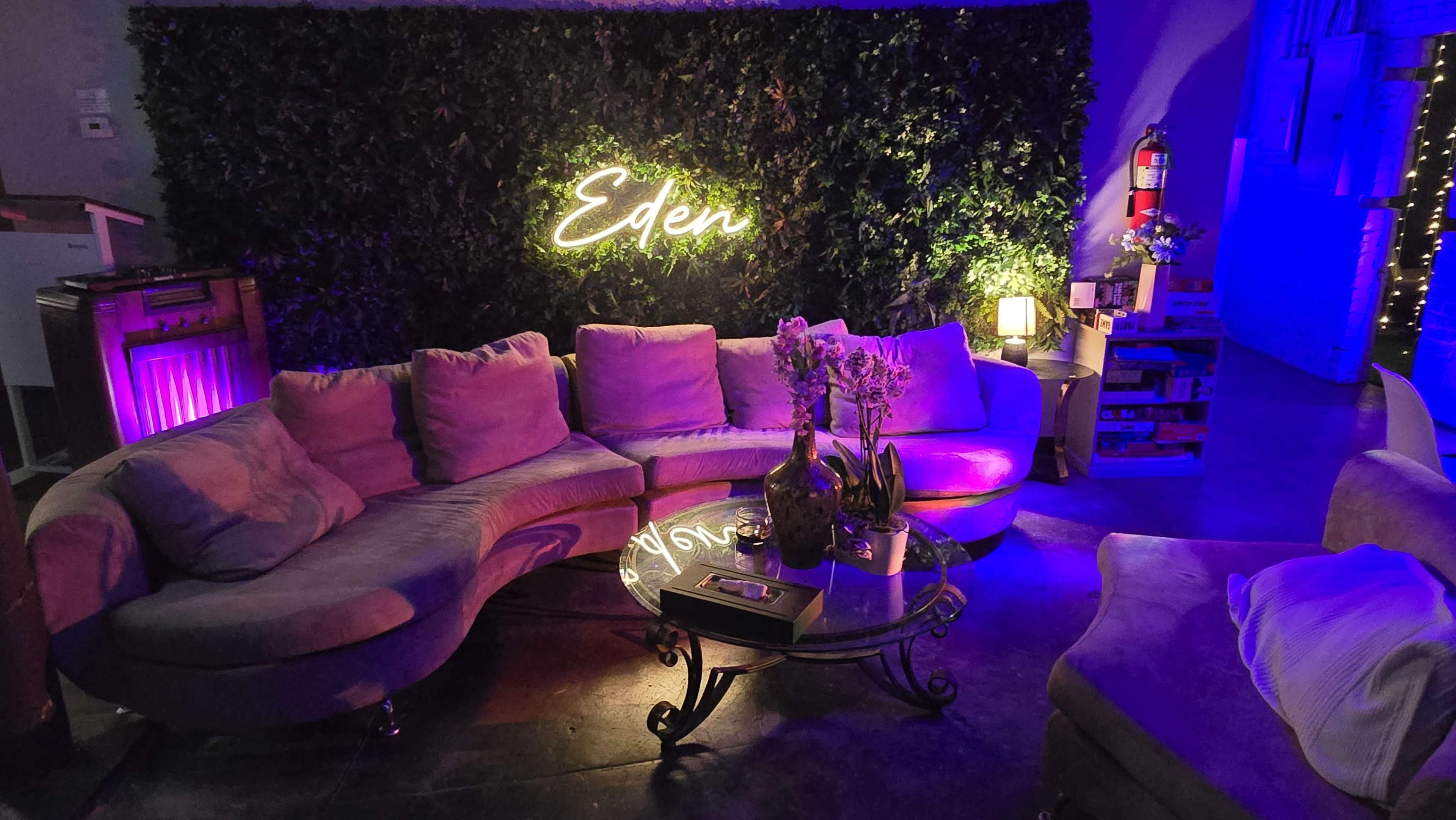 A curved sectional couch is positioned in front of a green wall with the neon word "Eden," surrounded by soft lighting and decorative plants.