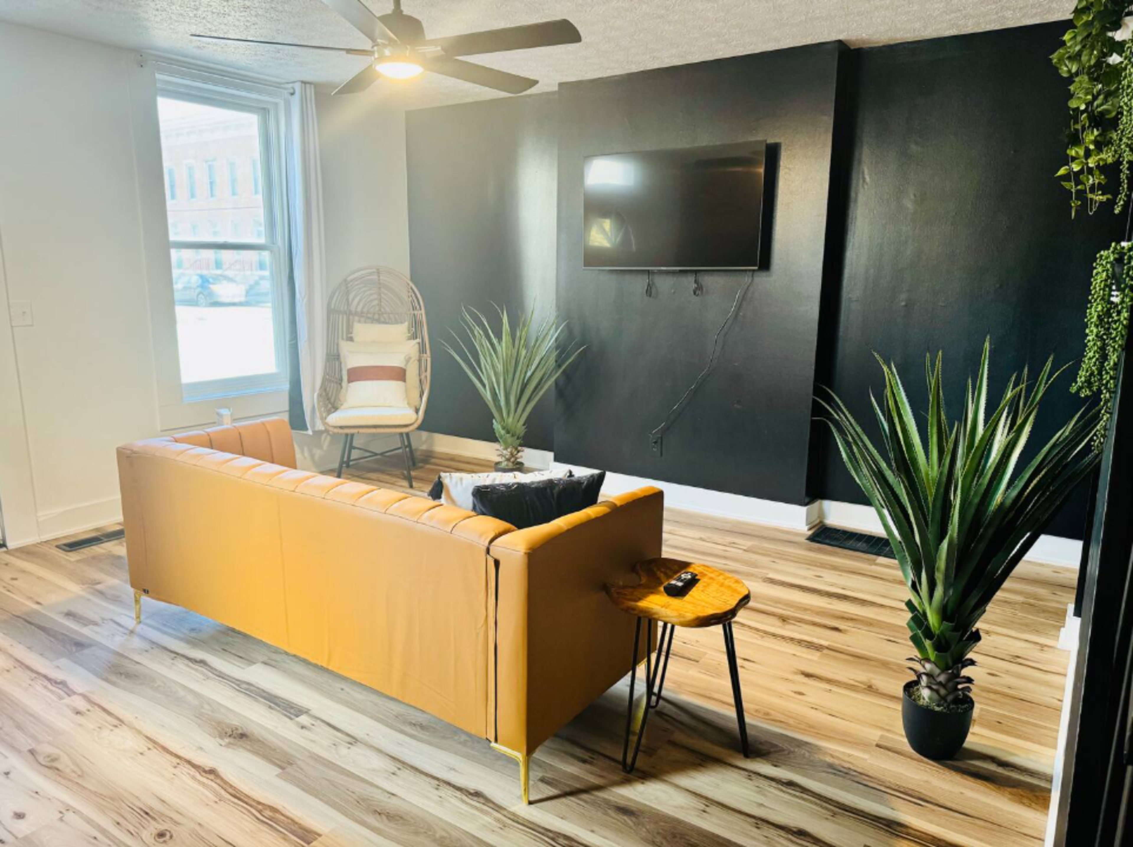 A modern living room features a tan leather couch, a small round table, a wall-mounted TV, and large planters with indoor plants.