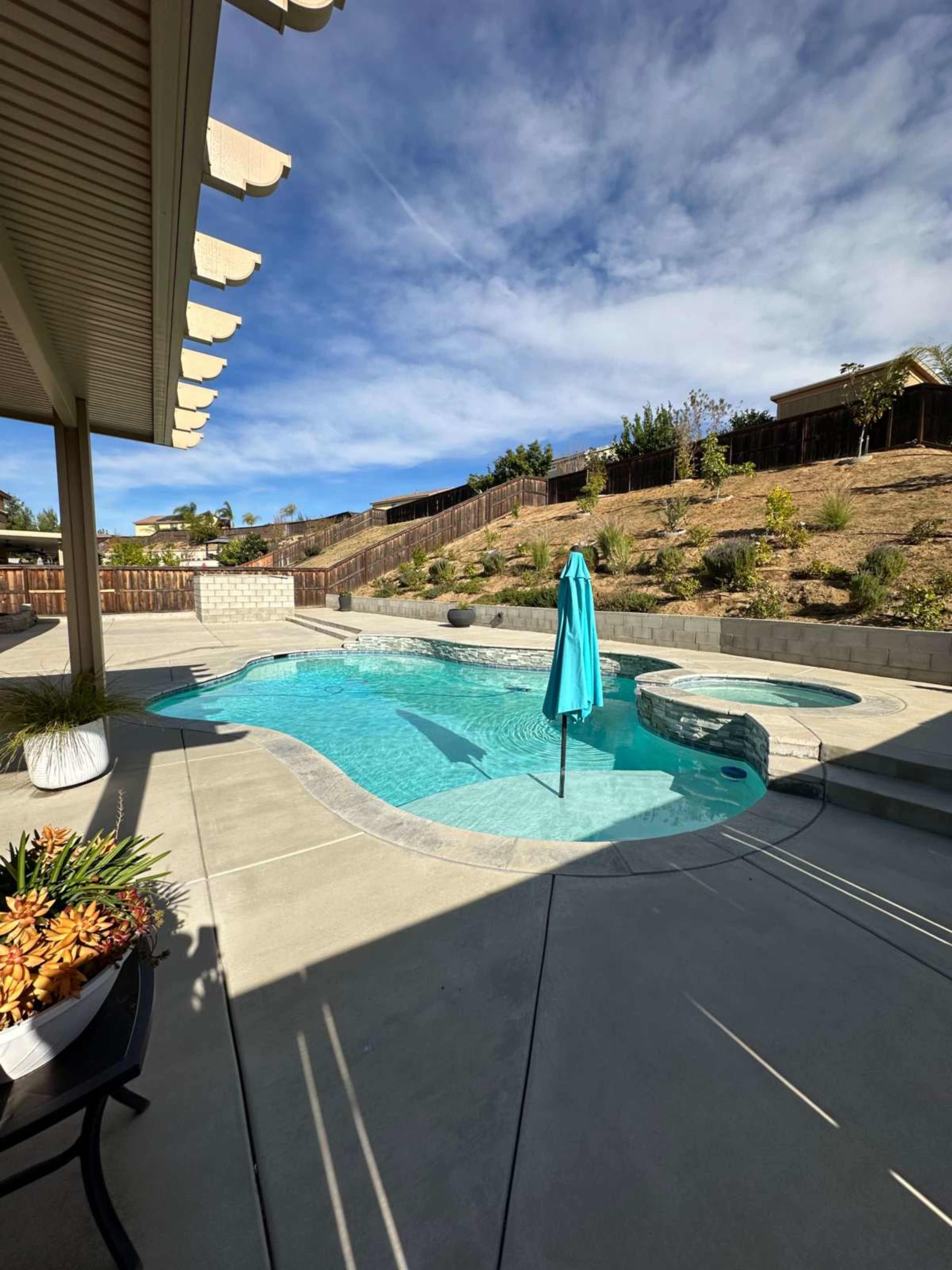 The image shows a backyard pool area with a turquoise umbrella, a spa section, and landscaped surroundings under a partly cloudy sky.