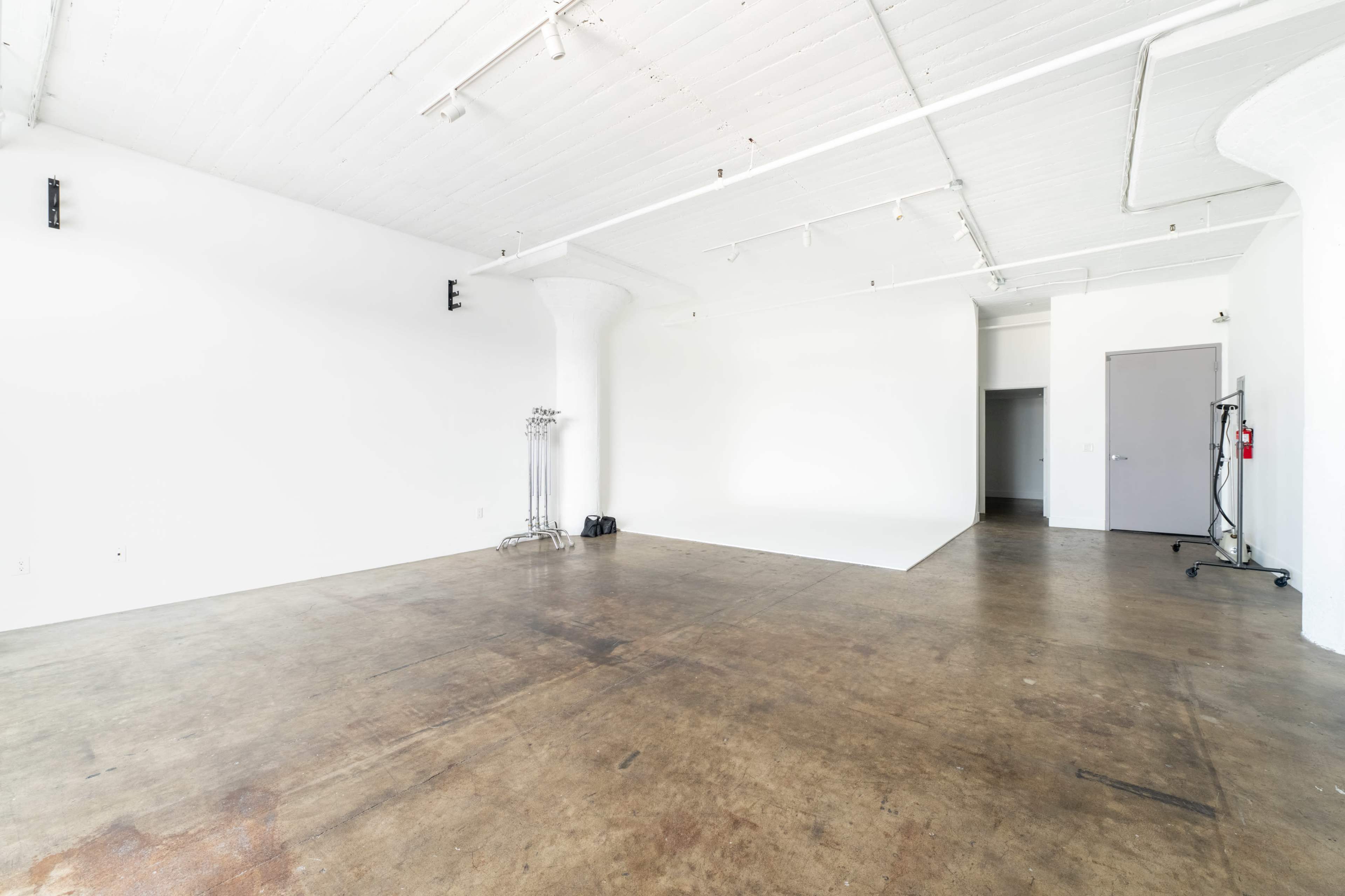 The image shows a spacious, empty studio with a white ceiling and walls, featuring concrete flooring and minimal furnishings.