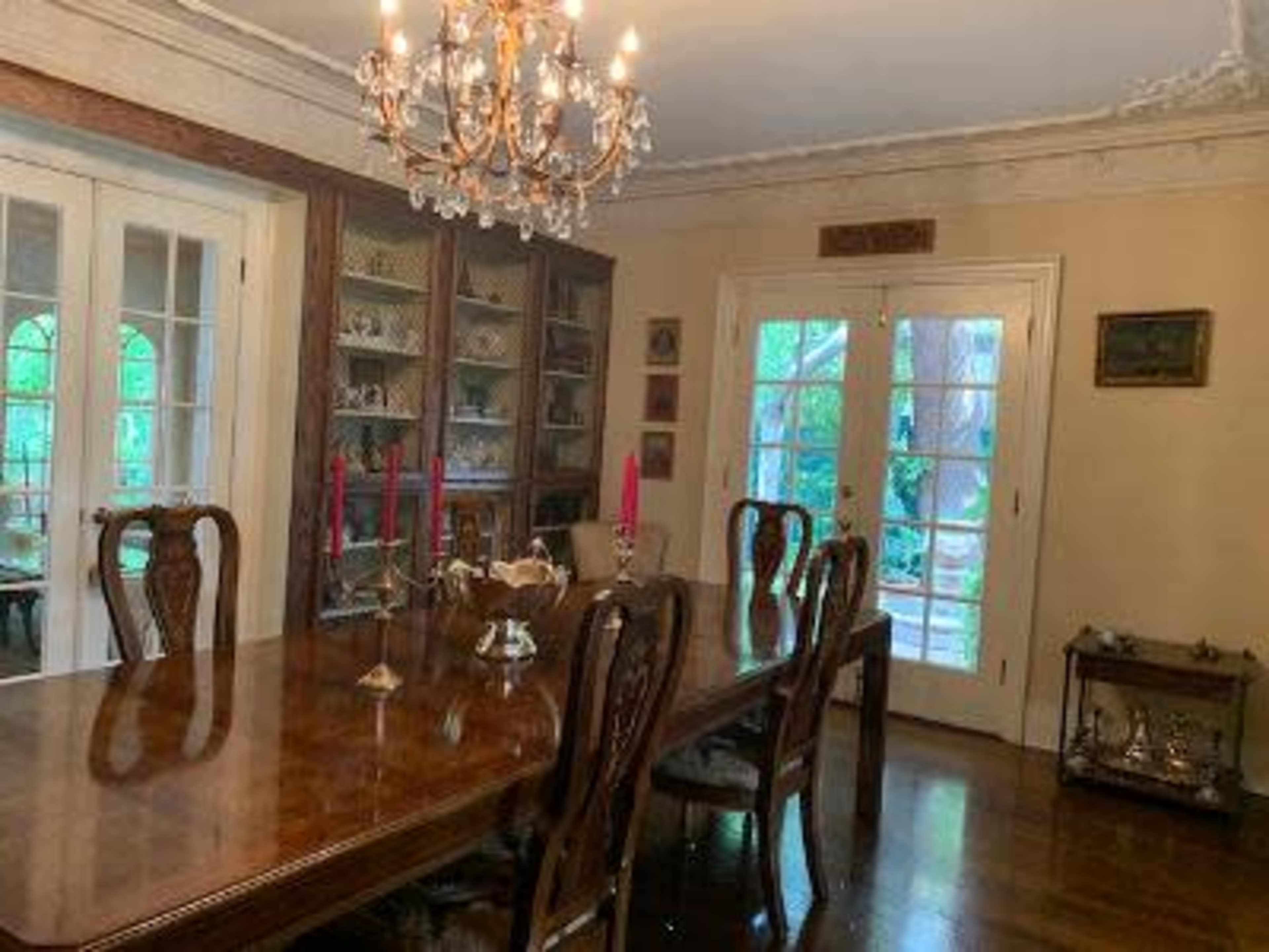 The image shows a dining room with a wooden table, several chairs, a chandelier, and a glass cabinet displaying decorative items.
