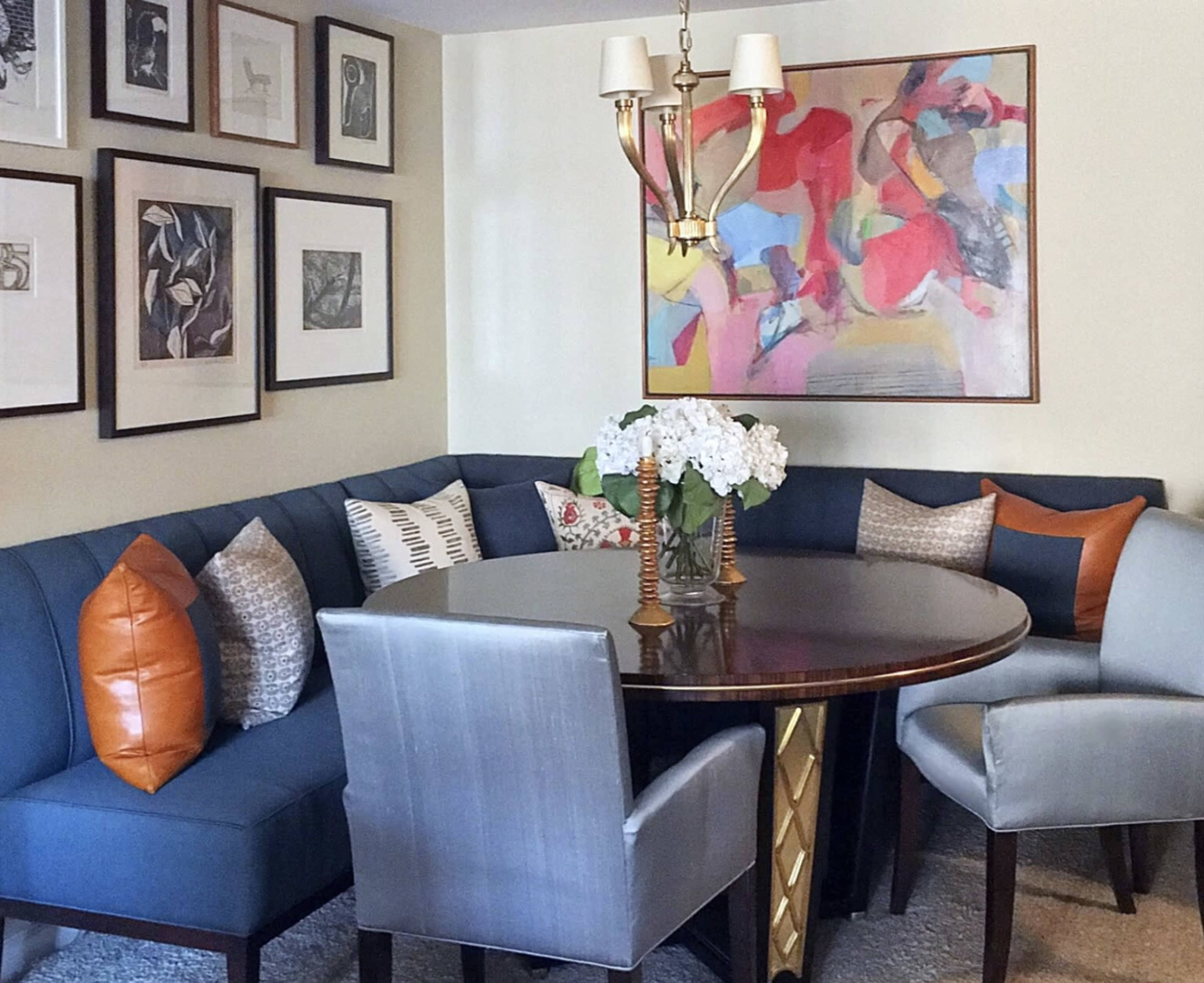 A circular table is surrounded by a blue upholstered banquette and light gray chairs, with framed artwork on the walls and a floral centerpiece on the table.